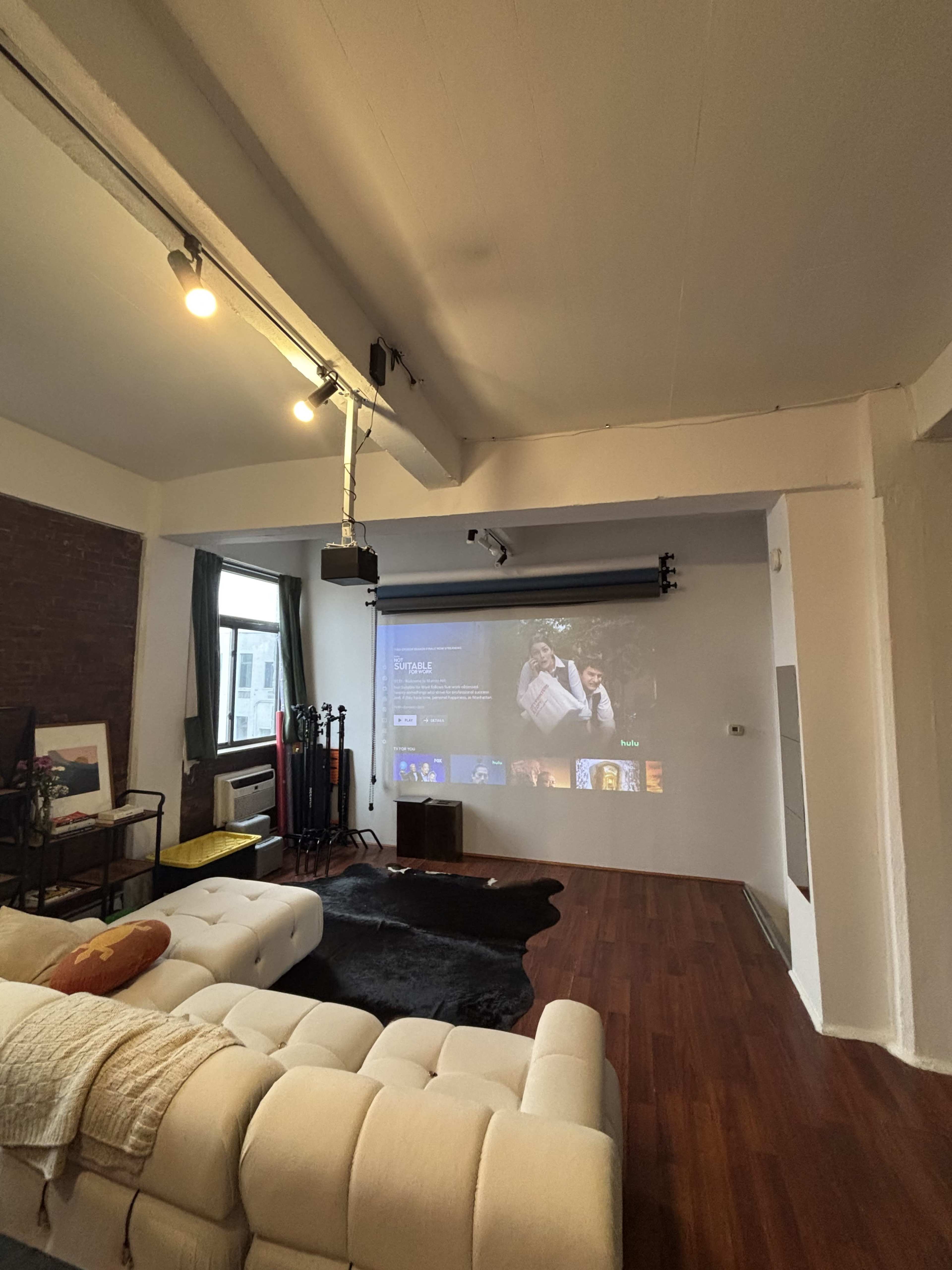 The image shows a modern living room with a white sectional sofa, a projector screen displaying a scene, and wooden flooring.