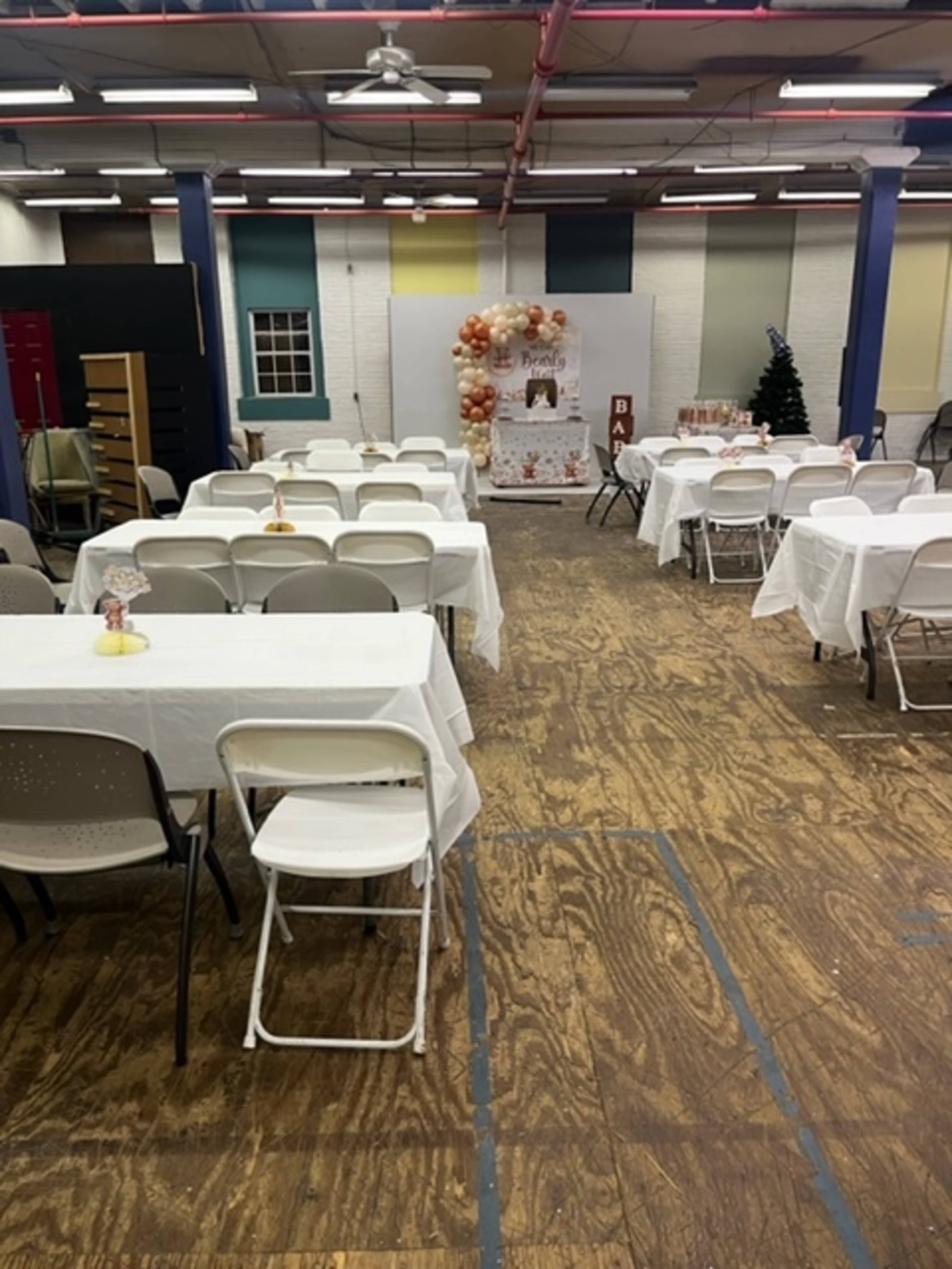 The image shows an indoor event space with several tables covered in white tablecloths, set up for an occasion, and festive decorations in the background.