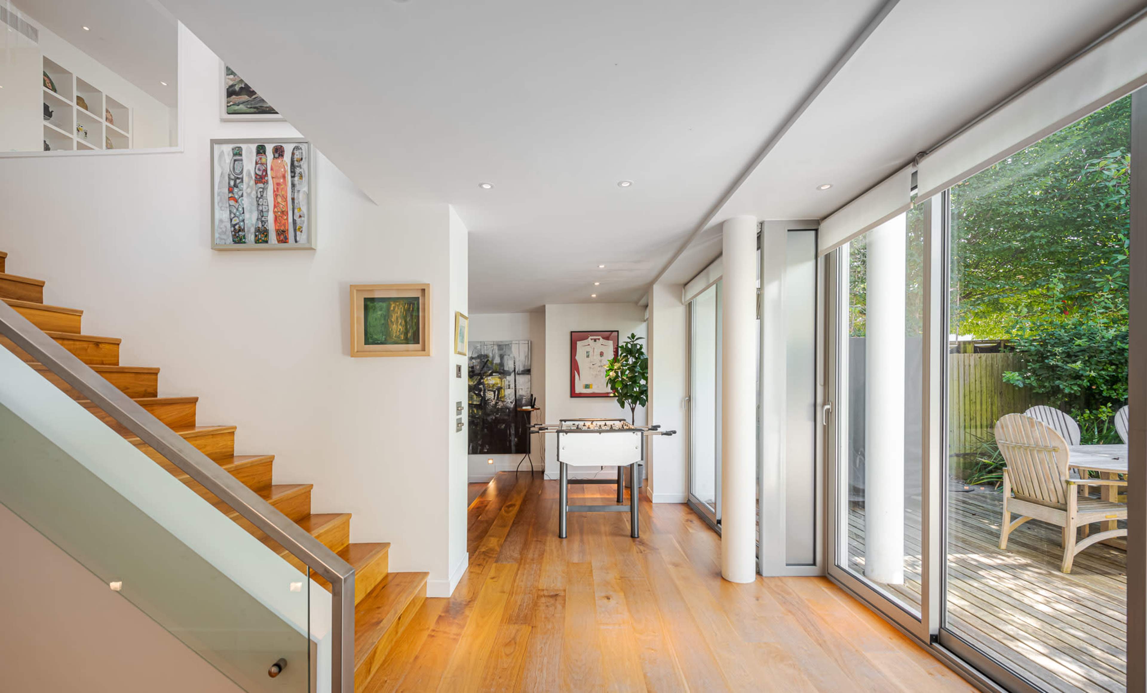 The image shows a modern interior with a wooden staircase leading to a bright open space featuring large sliding glass doors that open to a deck area.