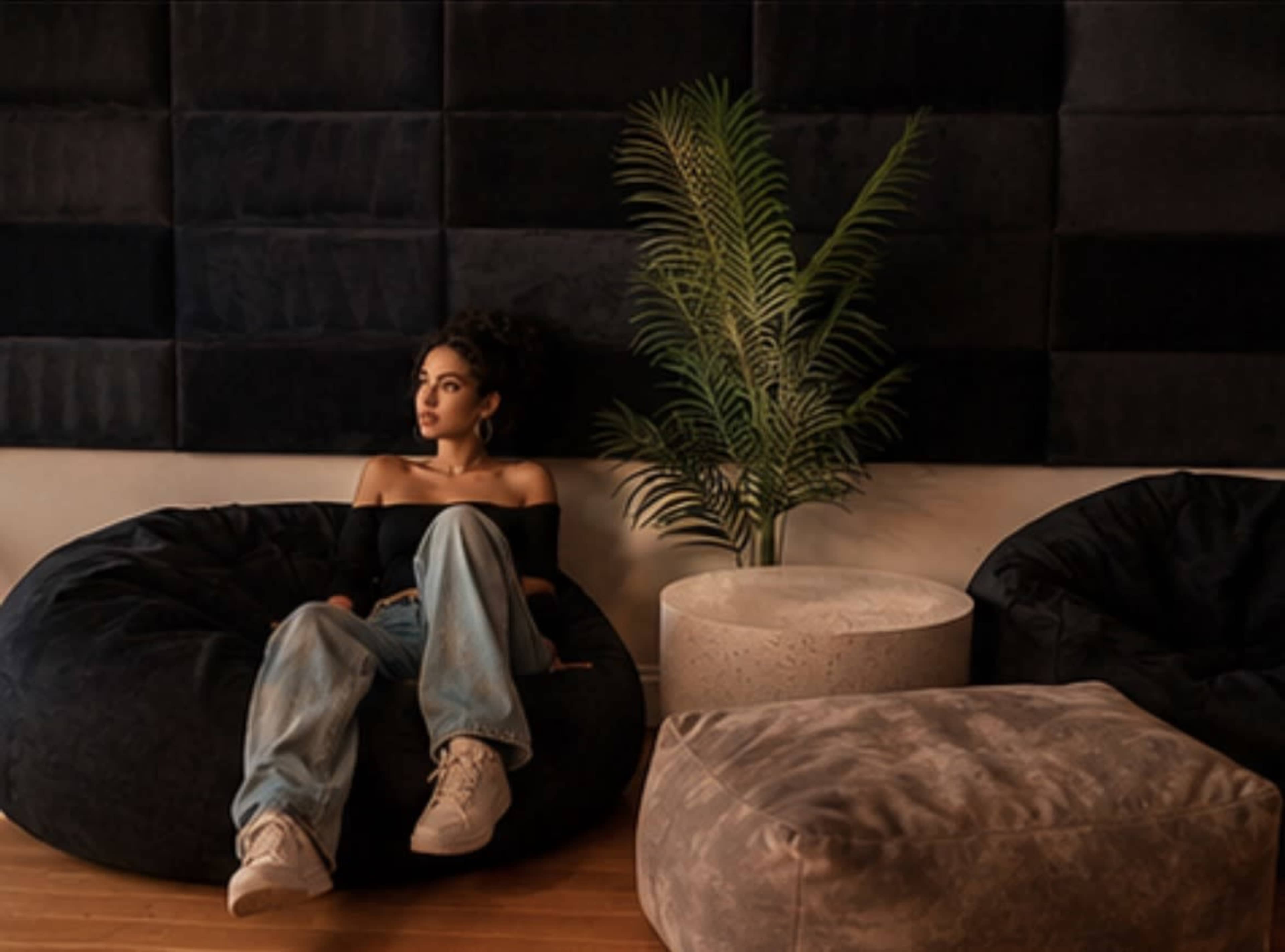 A woman sits on a bean bag chair in a modern room with dark wall panels and a potted plant beside a light-colored side table.