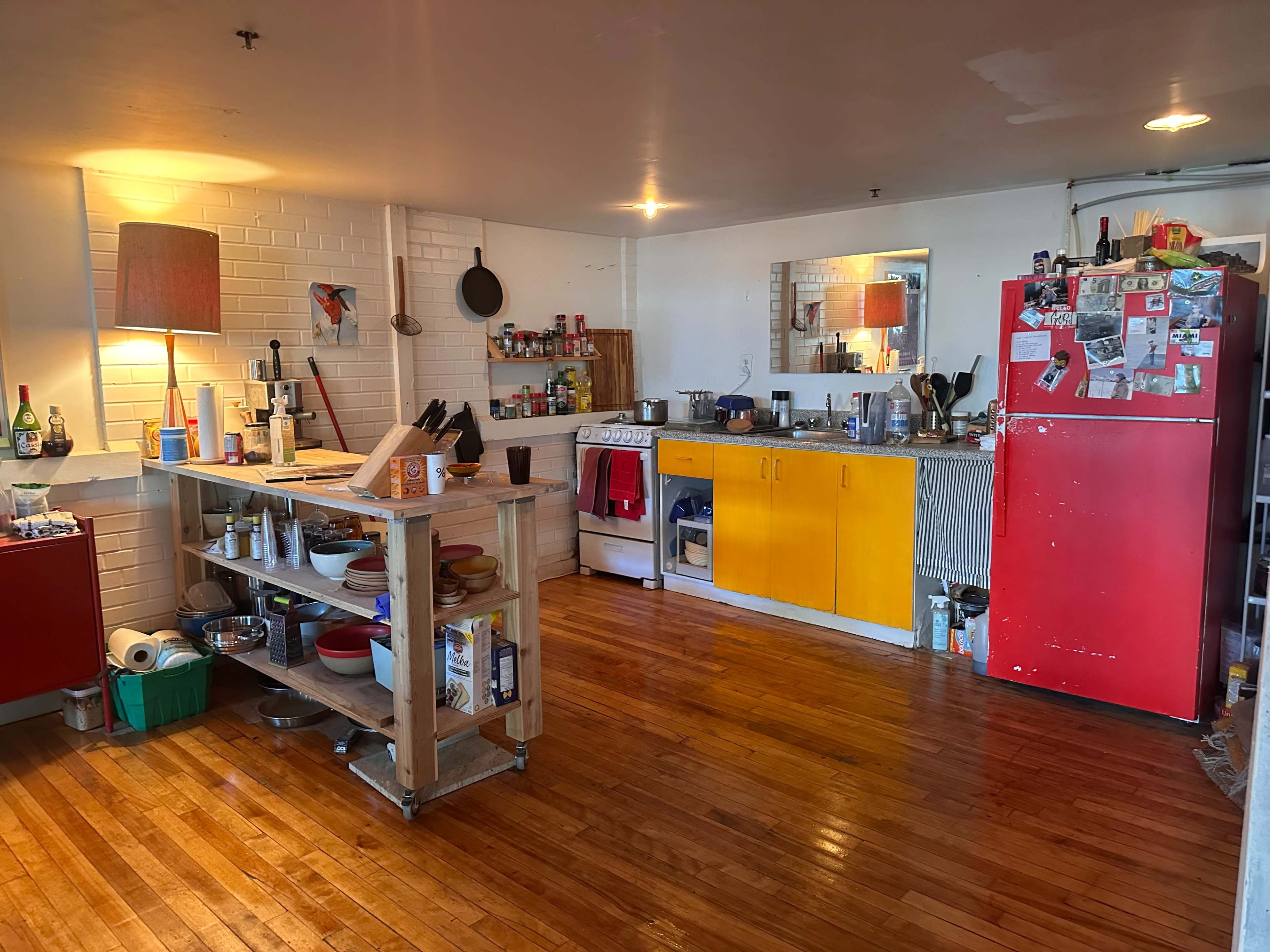 The image shows a spacious kitchen with wooden floors, featuring a yellow cabinet, a red refrigerator covered in papers, and various cooking utensils on a wooden island.