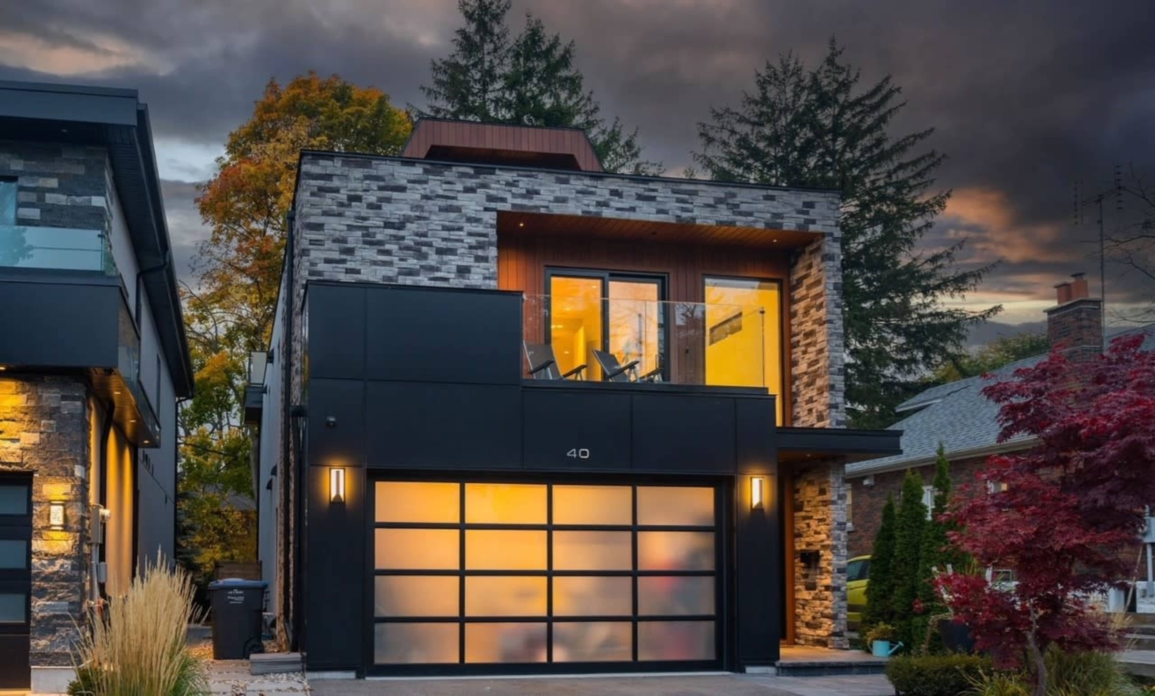 A modern two-story house with a stone and dark panel exterior, featuring a garage with frosted glass doors and a balcony on the upper floor.