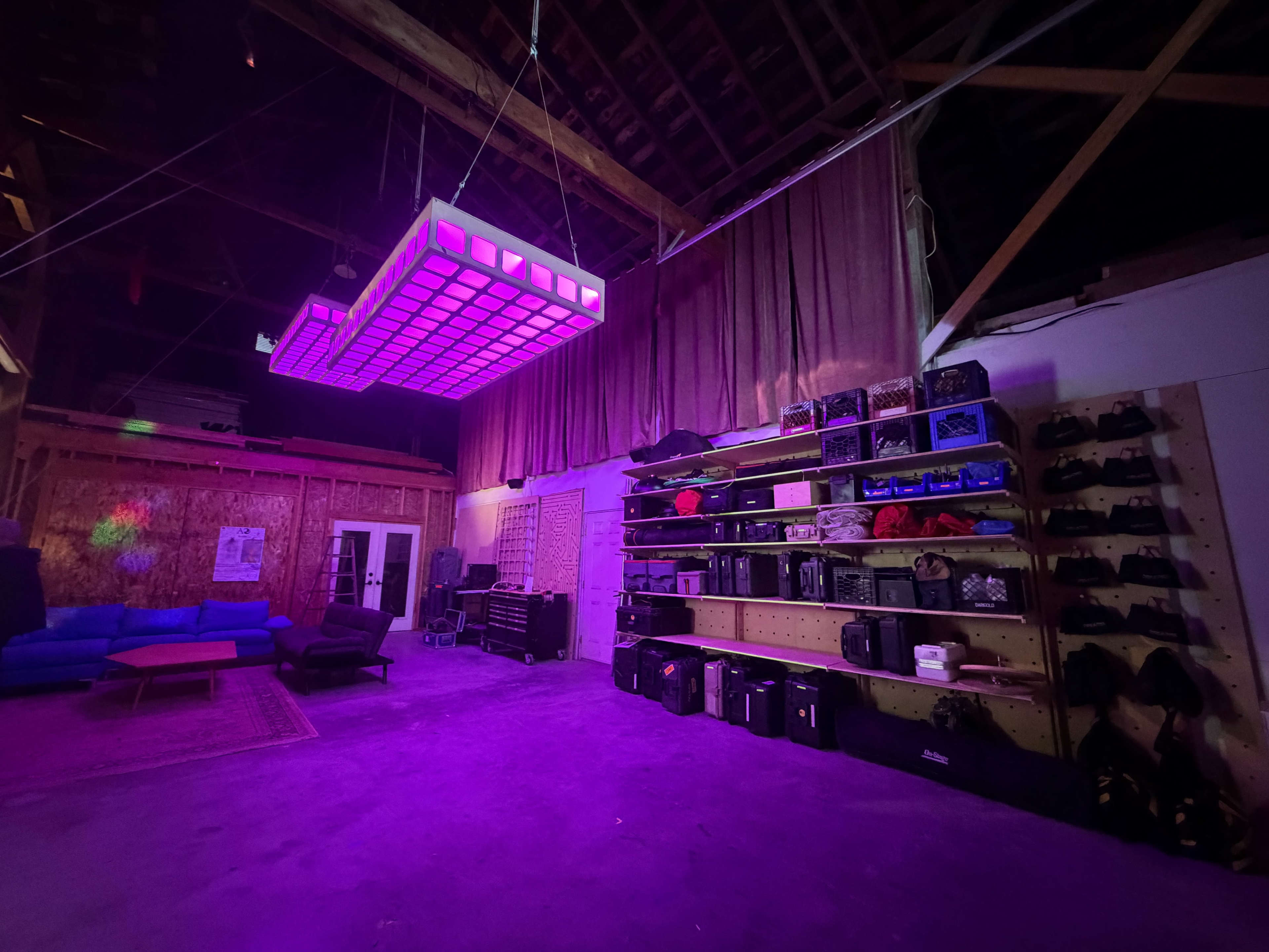 A large, illuminated LED panel hangs from the ceiling in a spacious warehouse, while shelves lined with various equipment and bags occupy one wall.