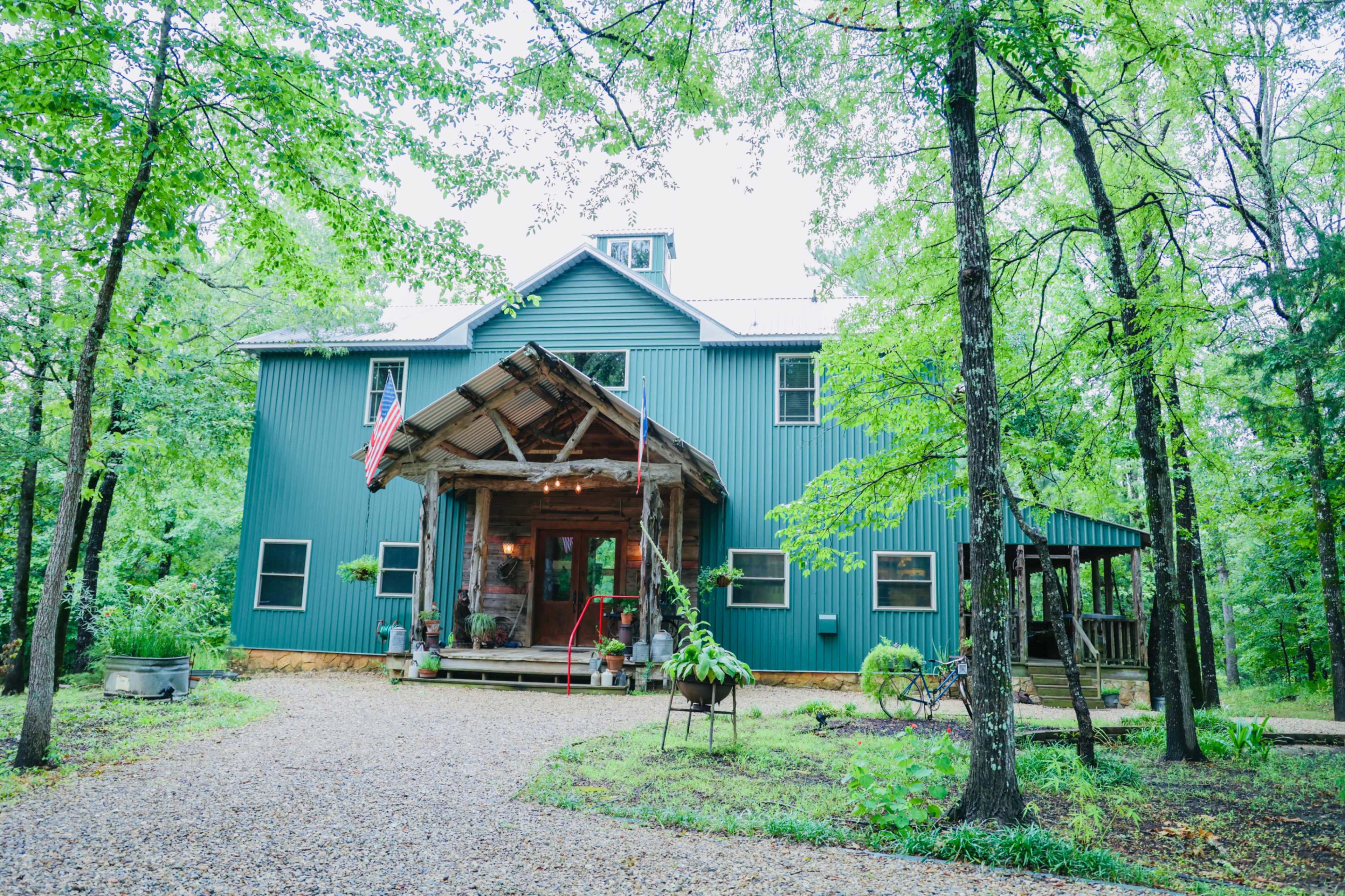 A large blue house with a wooden porch and an American flag stands surrounded by trees and a gravel driveway.