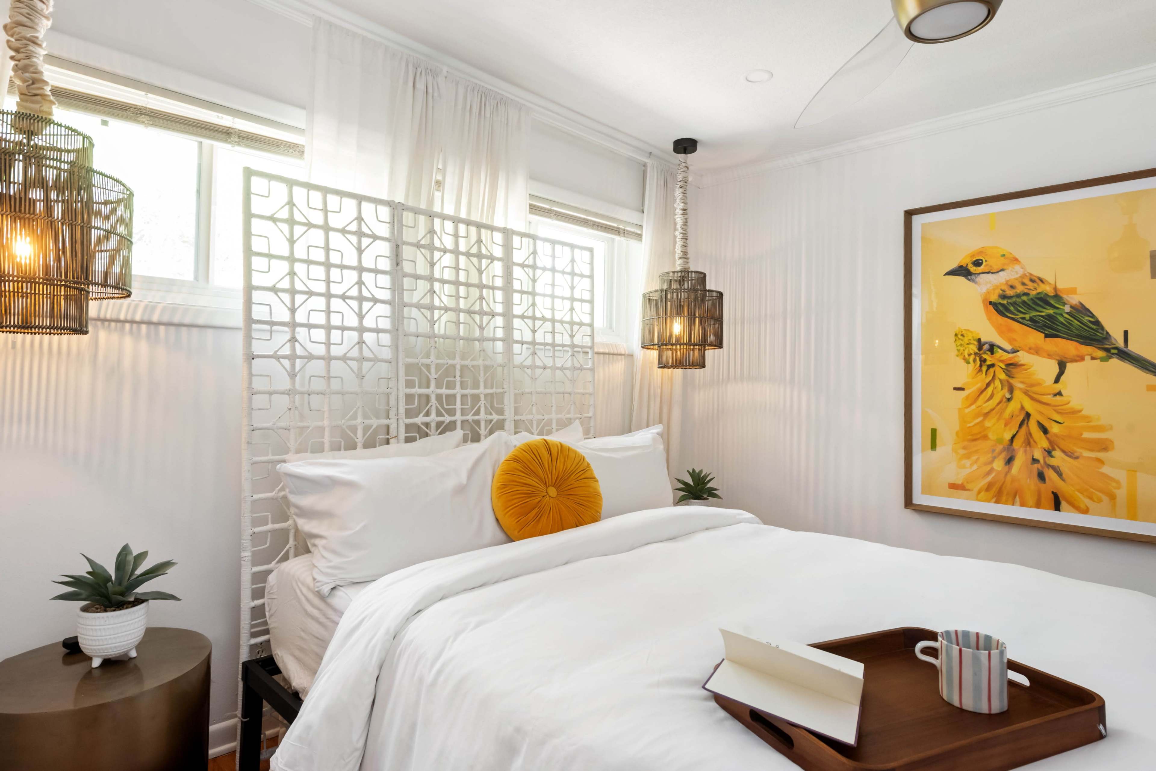 The image shows a cozy bedroom with a bed adorned with white linens, a decorative yellow pillow, a wooden tray with a mug, and artwork featuring a bird on the wall, illuminated by pendant lighting.