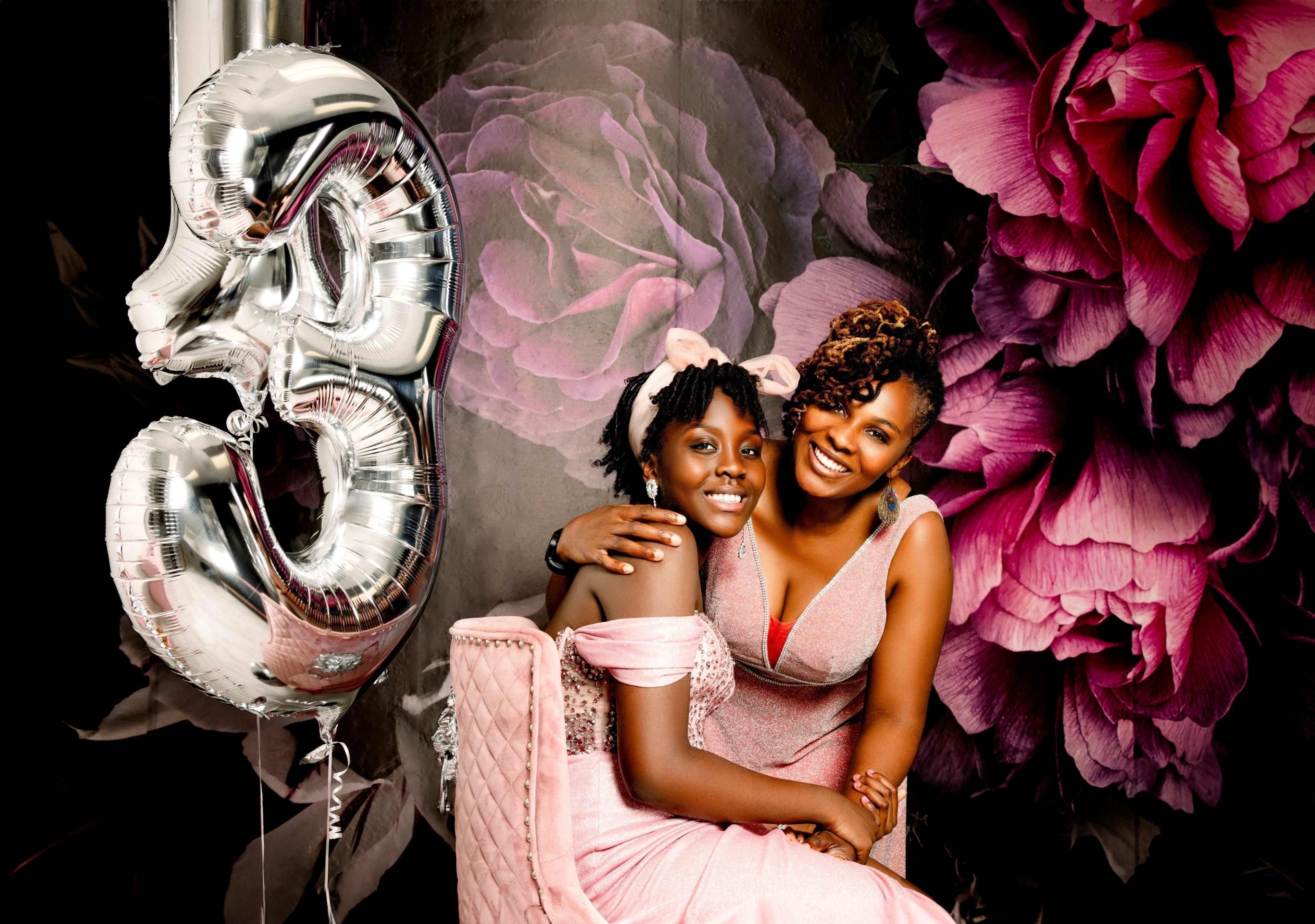 A woman and a girl pose together with a large silver "3" balloon in front of a floral backdrop.