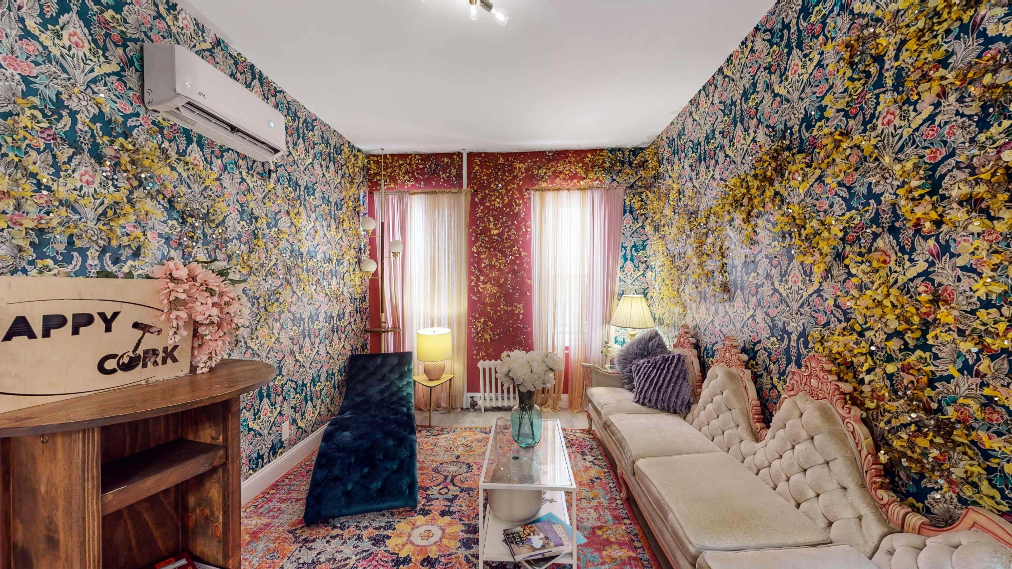 The room features floral wallpaper, a patterned area rug, a vintage sofa, and large windows with sheer curtains.
