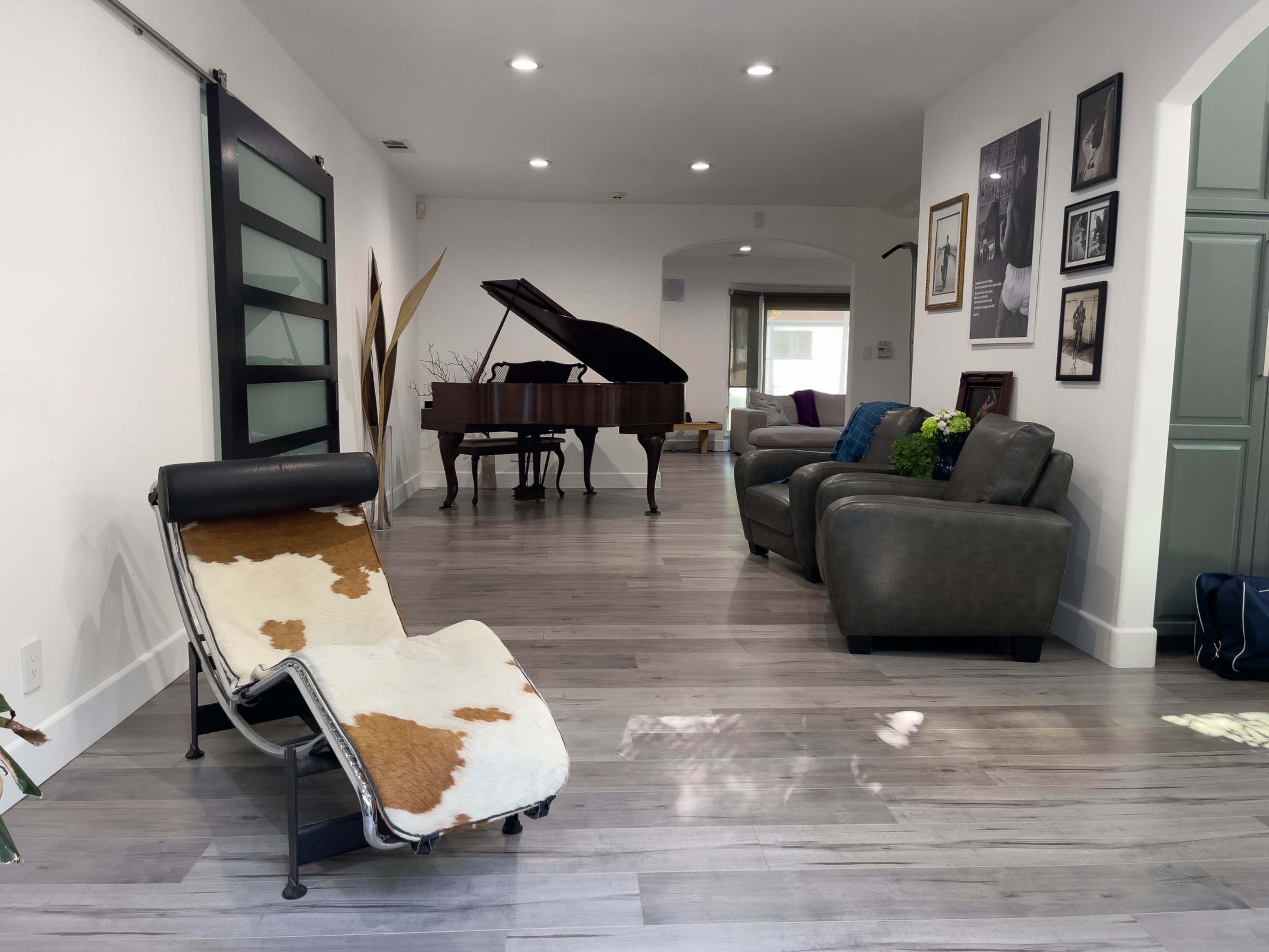 A spacious living area features a grand piano, a cowhide chair, and gray sofas along with various framed photographs on the walls.
