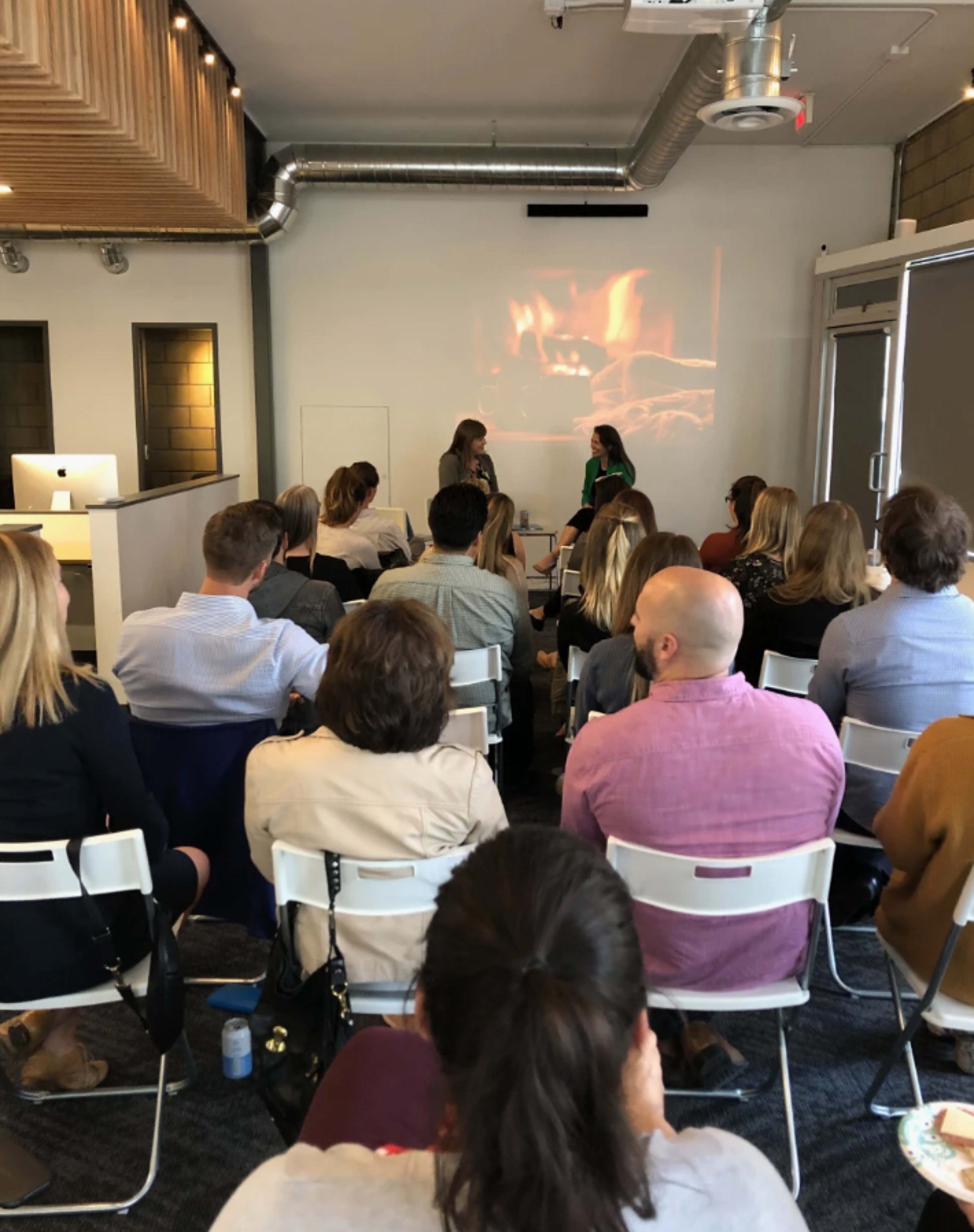 A group of people is seated in a conference room facing two women engaged in a discussion, with a projected image of a fireplace displayed behind them.