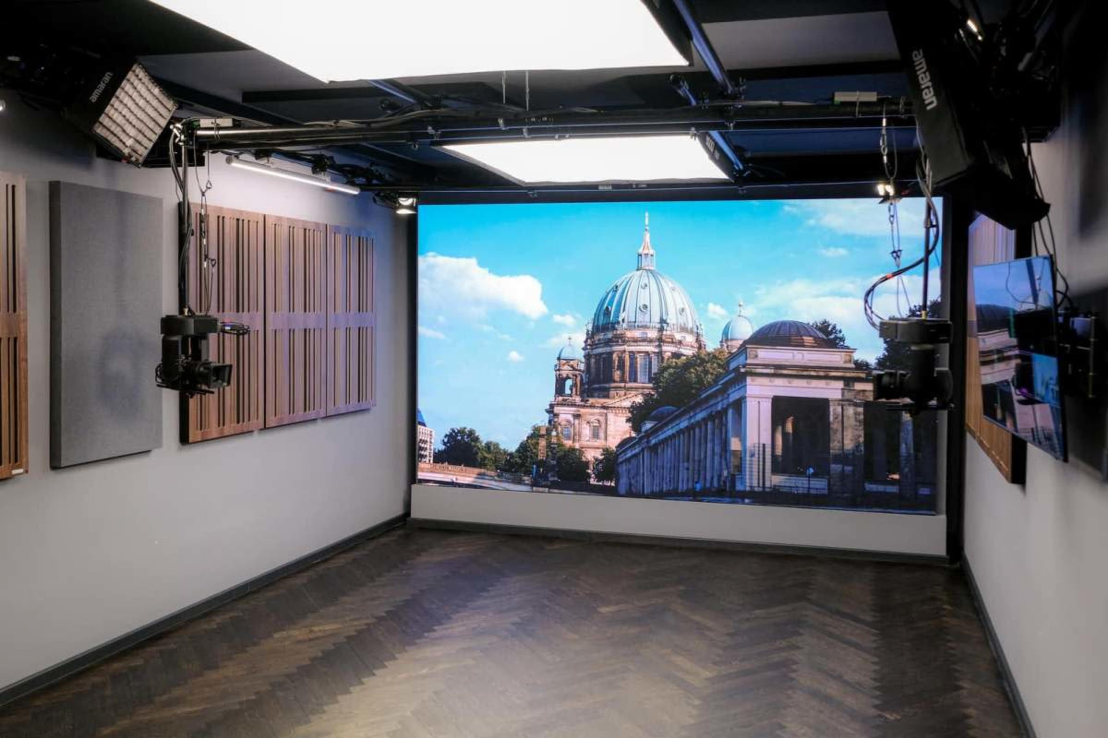 The image shows a modern studio setup with a large screen displaying a view of a building featuring a dome and columns, framed by a well-lit interior.