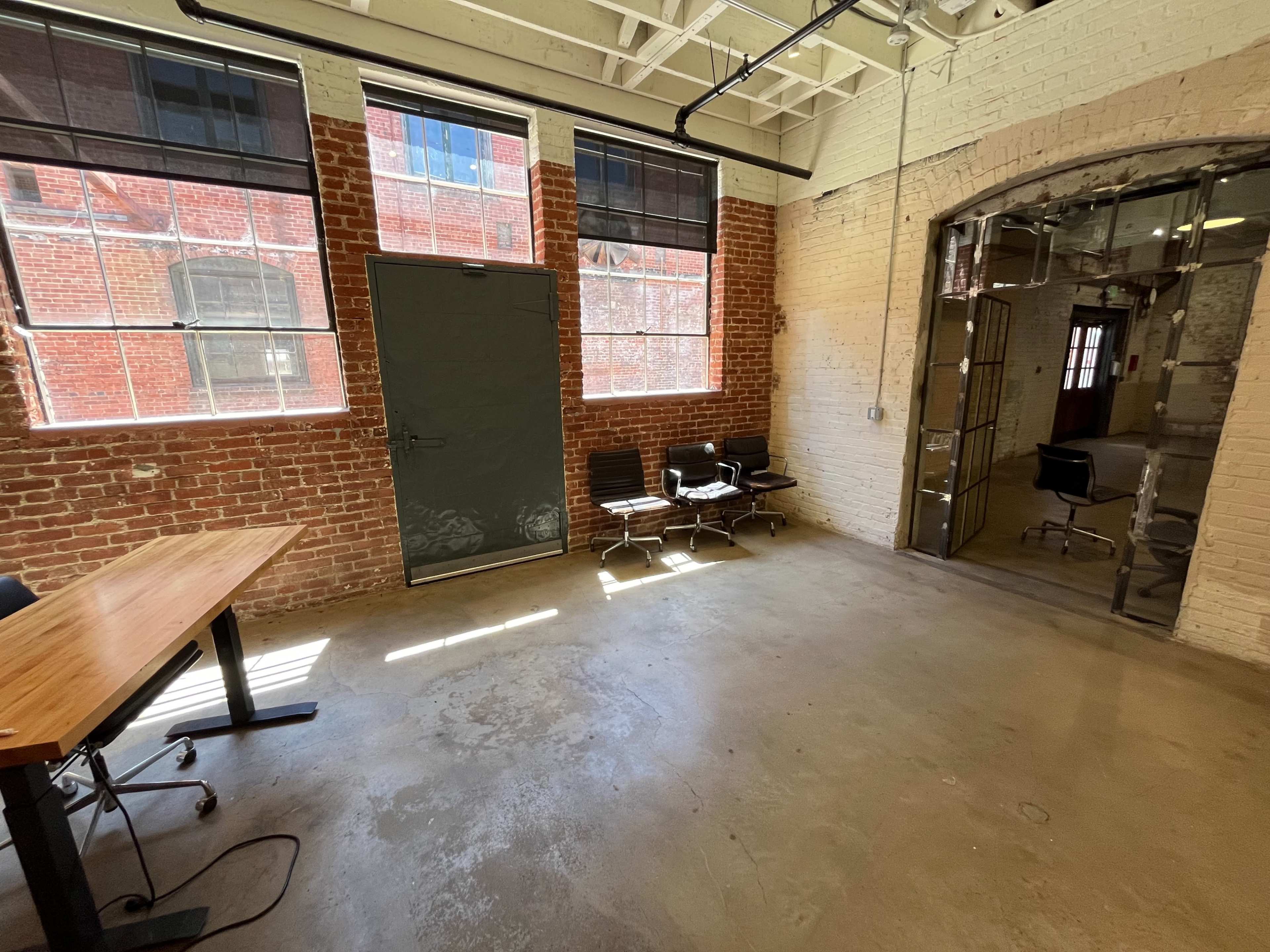 A minimalist office space with exposed brick walls, a wooden table, and several office chairs near a door and large windows allowing natural light in.