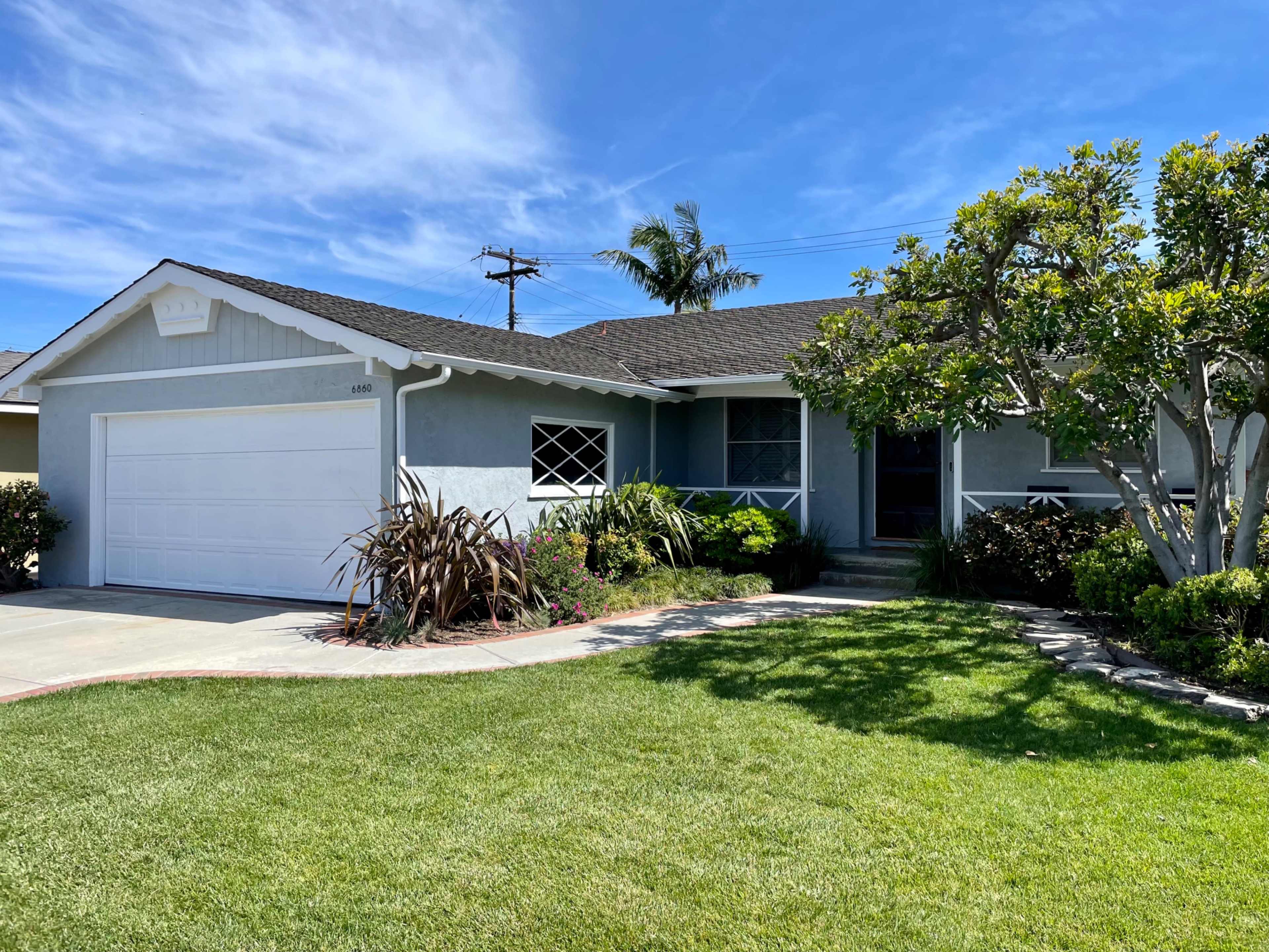 A single-story blue house with a white garage door and landscaped front yard is shown under a clear sky.