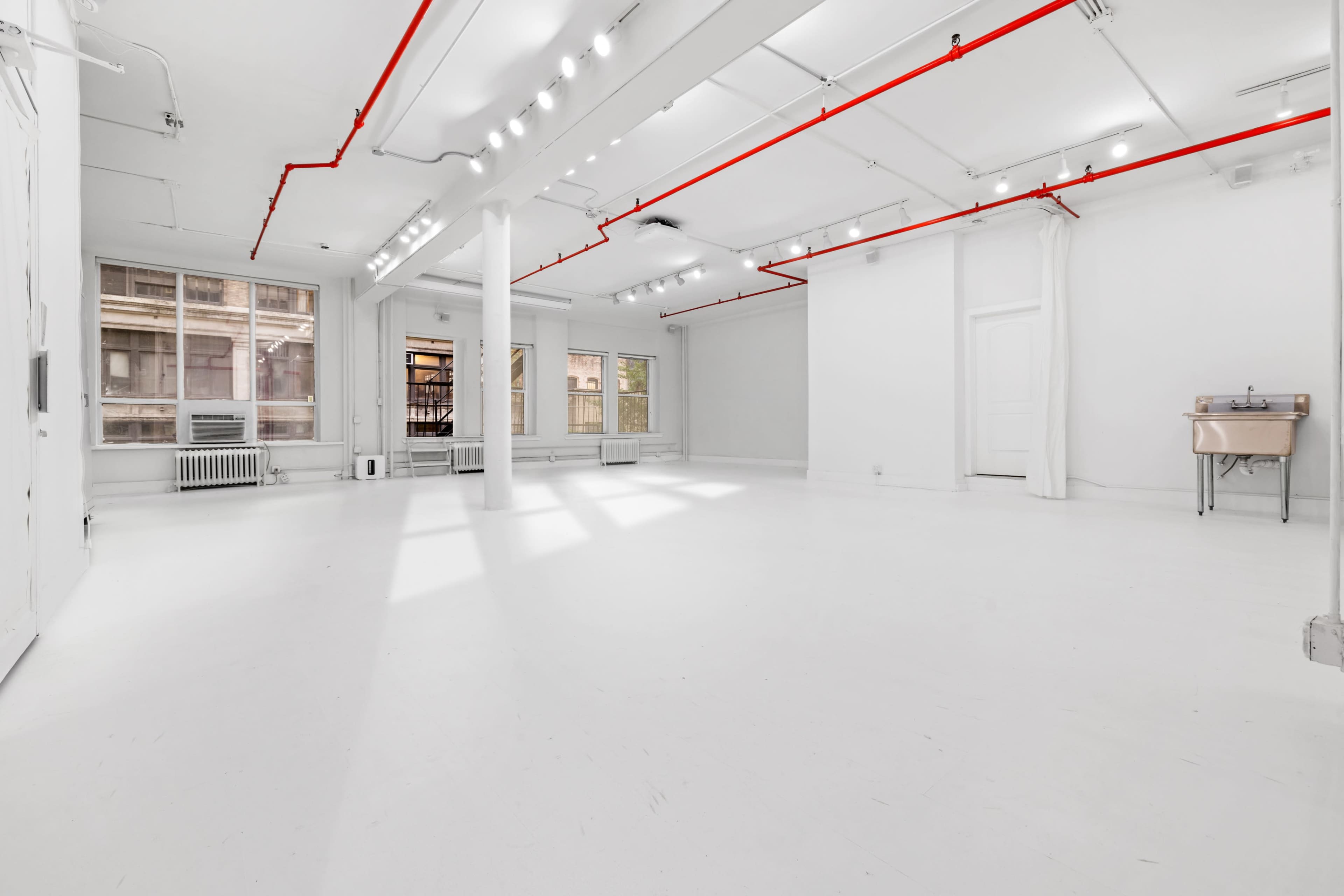 An empty, bright white room with large windows and red pipes along the ceiling.