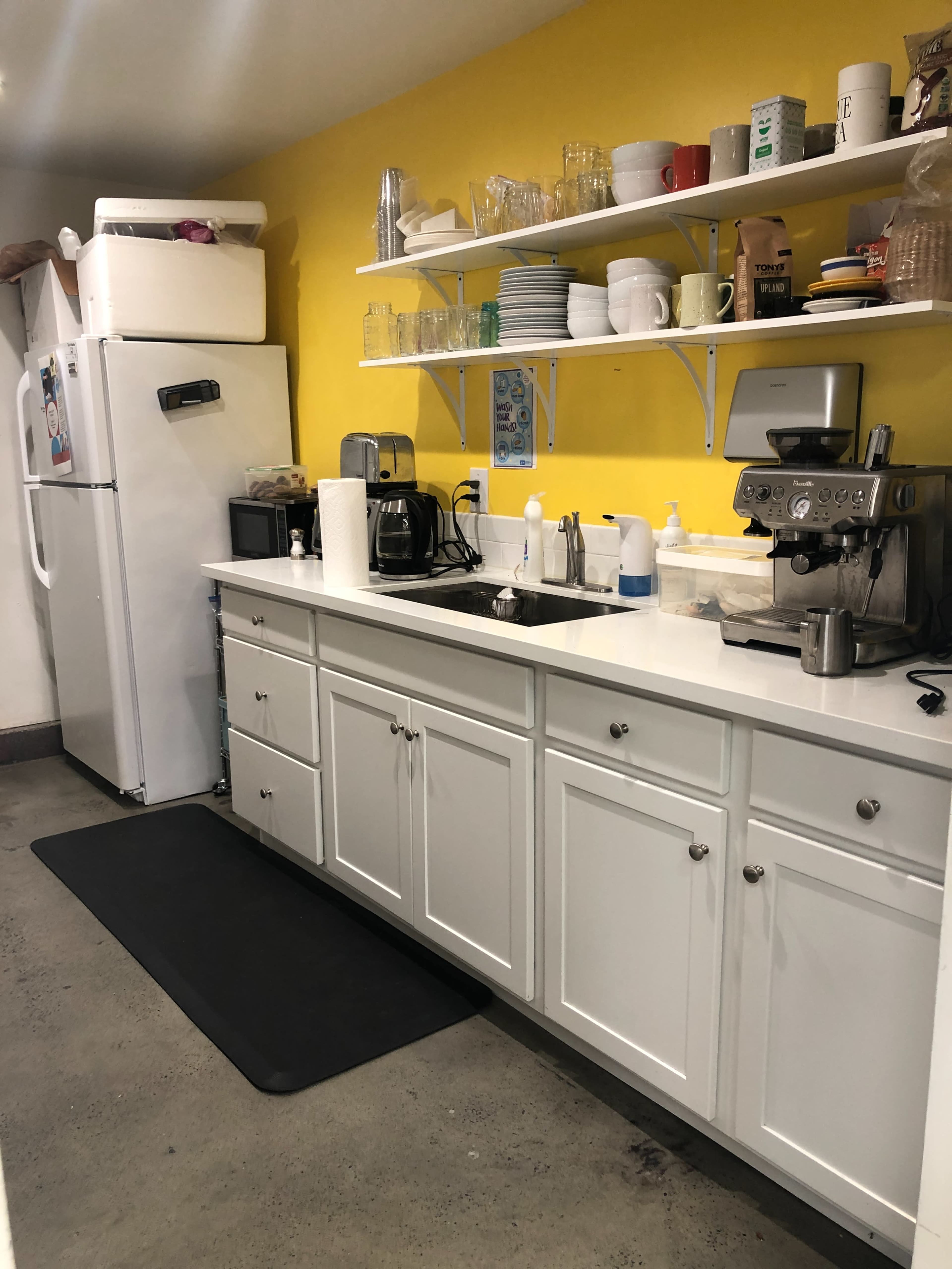 A kitchen with white cabinets, a yellow wall, and various appliances, including a refrigerator, coffee machine, and shelves holding dishes and glasses.