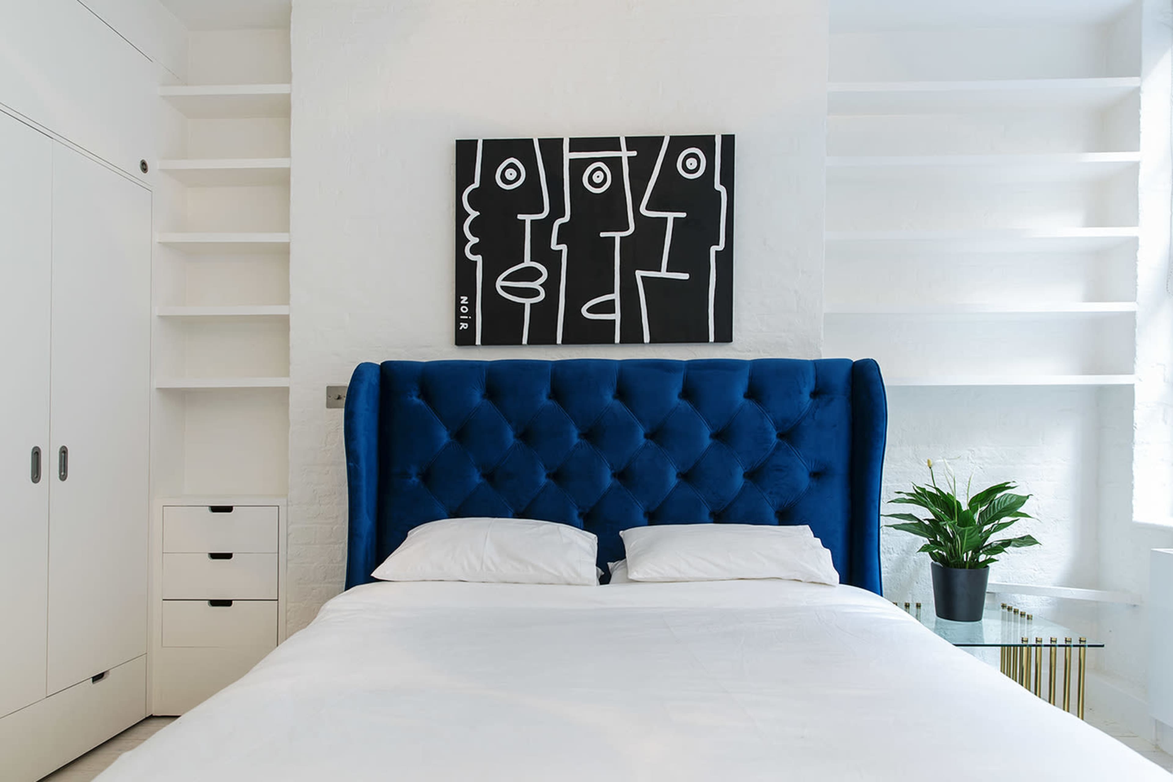 The image shows a bedroom with a large blue tufted headboard, a white bedding set, a black and white artwork above the bed, and minimal shelving and storage on the walls.