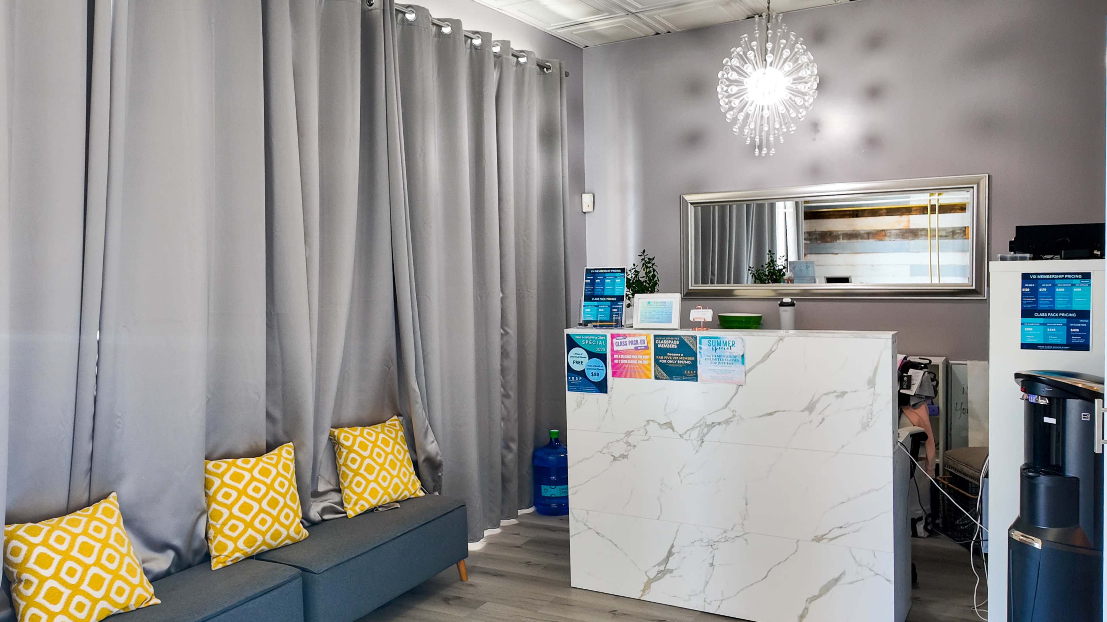 The image shows a reception area with gray curtains, a marble front desk, a mirror, a chandelier, and a couch with yellow patterned cushions.