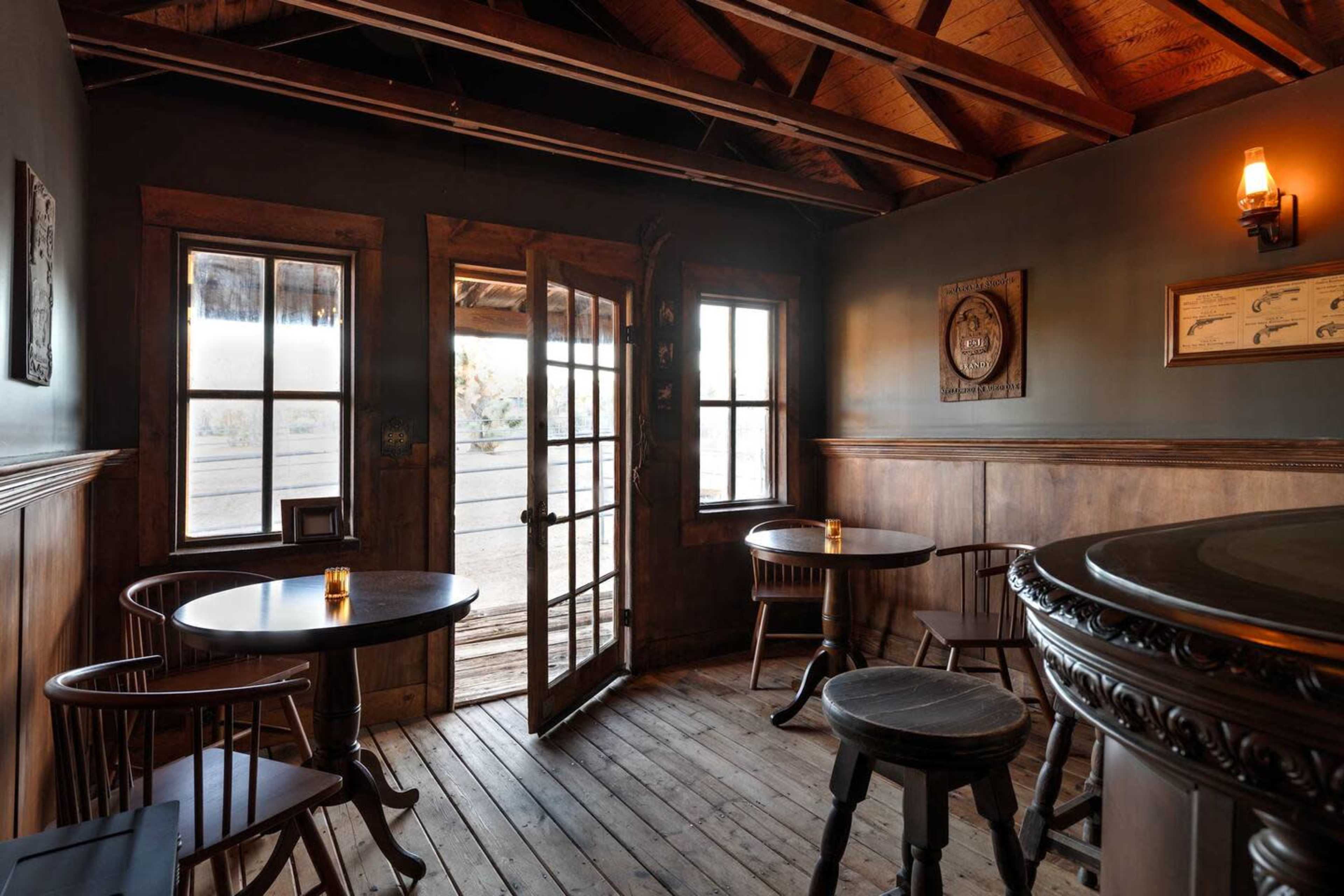The interior of a rustic tavern features wooden tables, a bar, and large windows that open to an outdoor view.