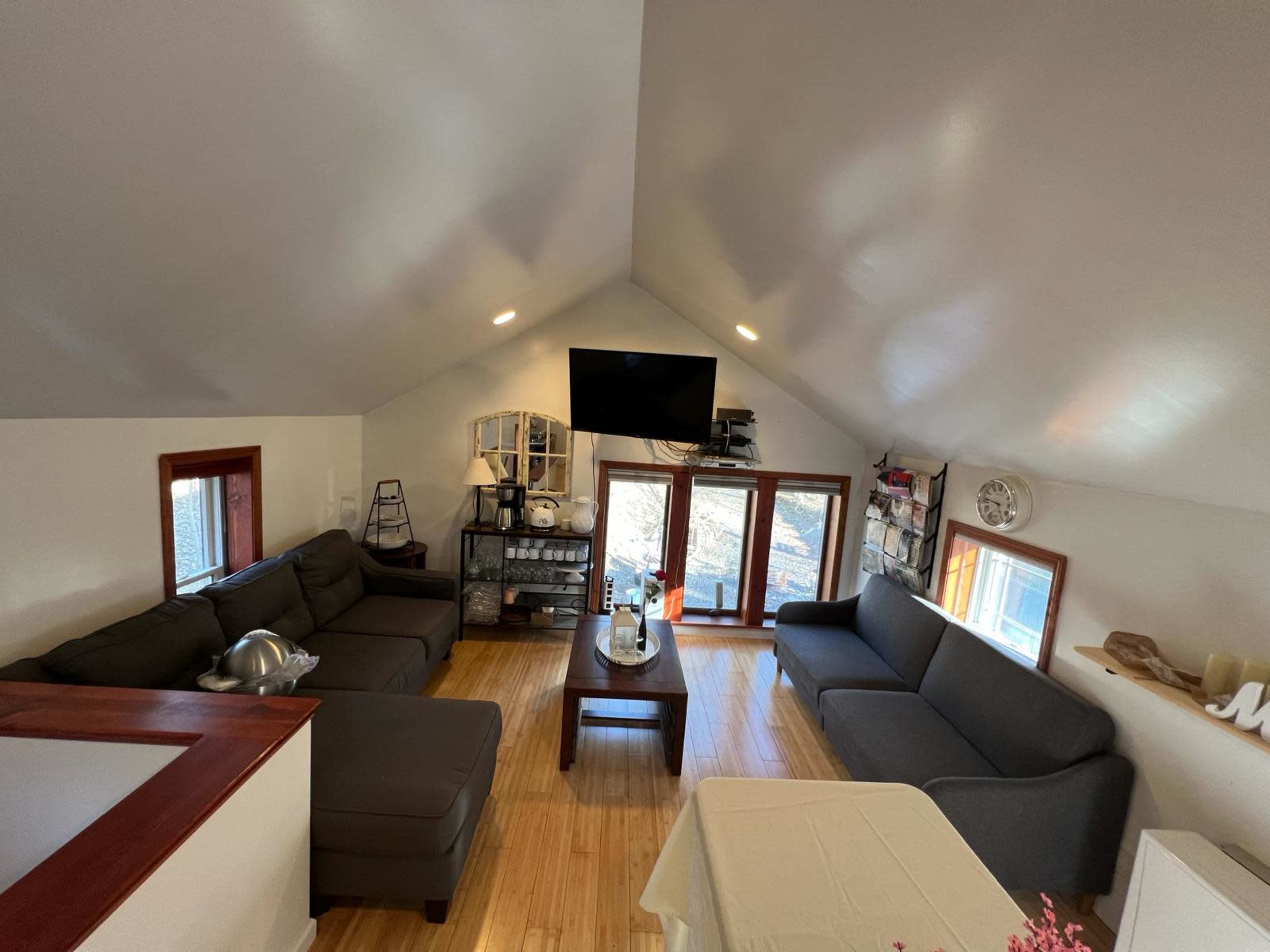 A spacious living room with two gray sofas, a television mounted on the wall, and large windows offering a view of the outdoors.