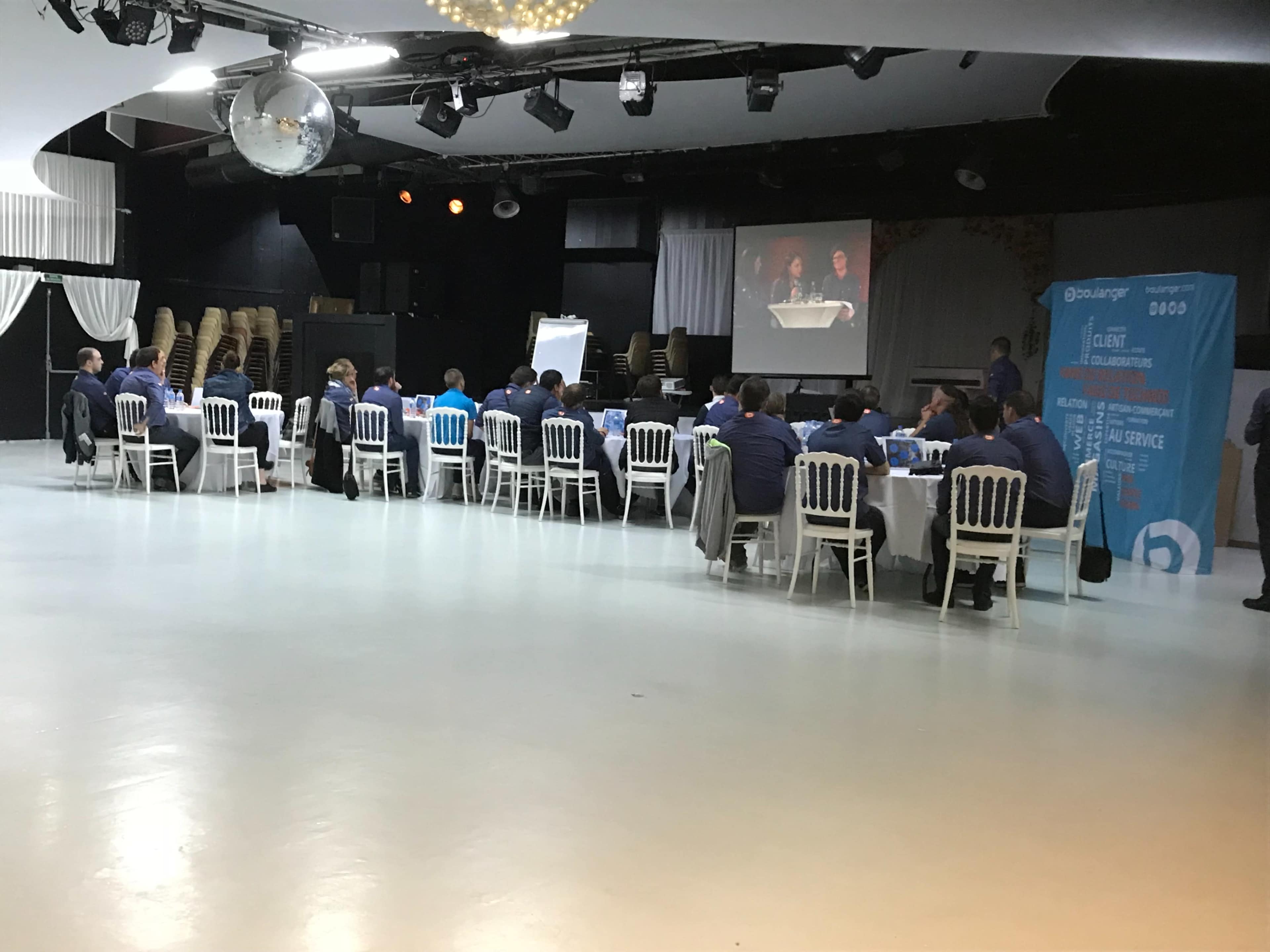 A group of people sits at a long table in a conference room, with a projector displaying a presentation on a screen.