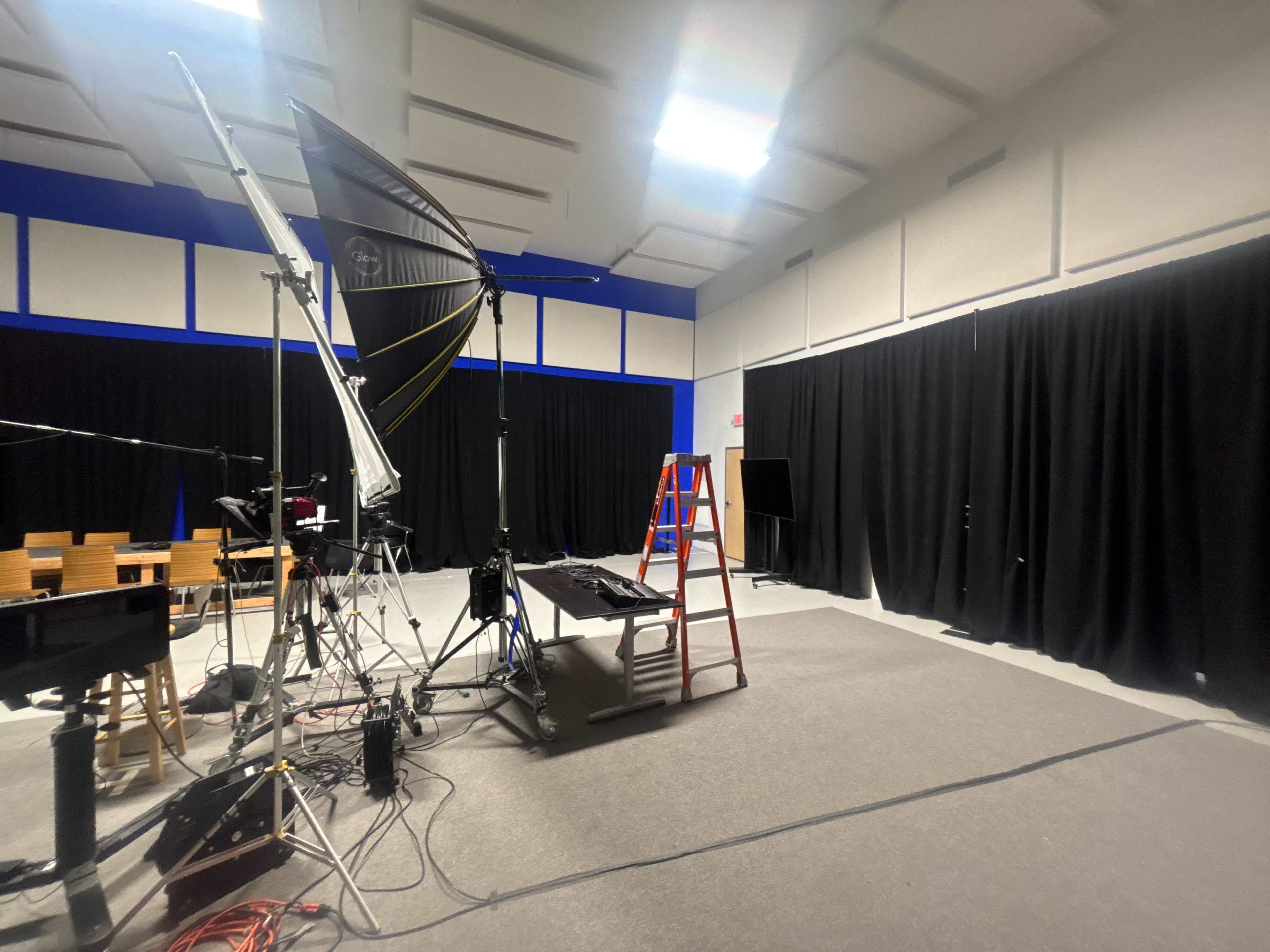 The image shows a studio space equipped with lighting equipment, a ladder, and black curtains, set up for a production or recording.