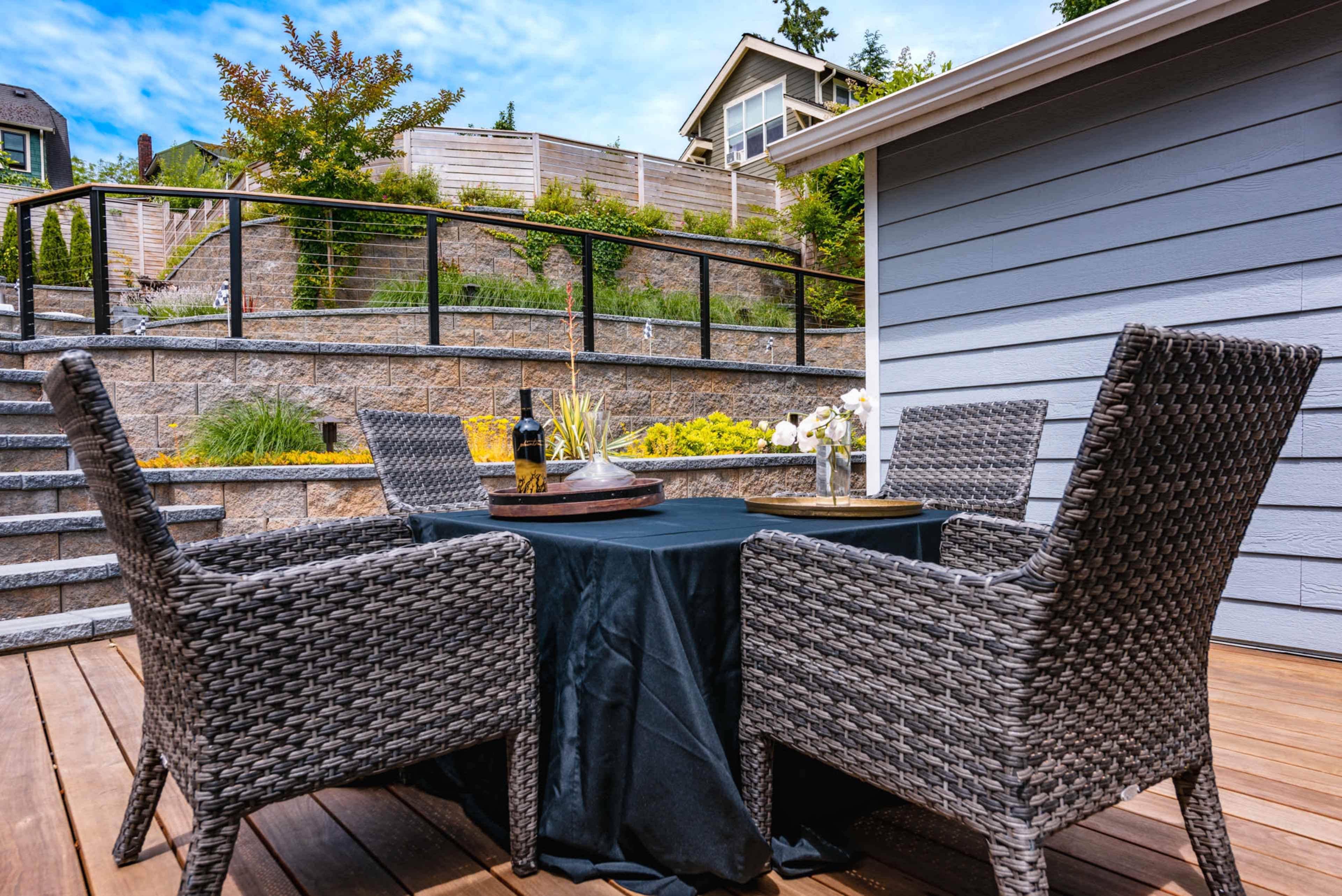 A dining table with two wicker chairs is set on a wooden deck overlooking landscaped stairs and a house in the background.