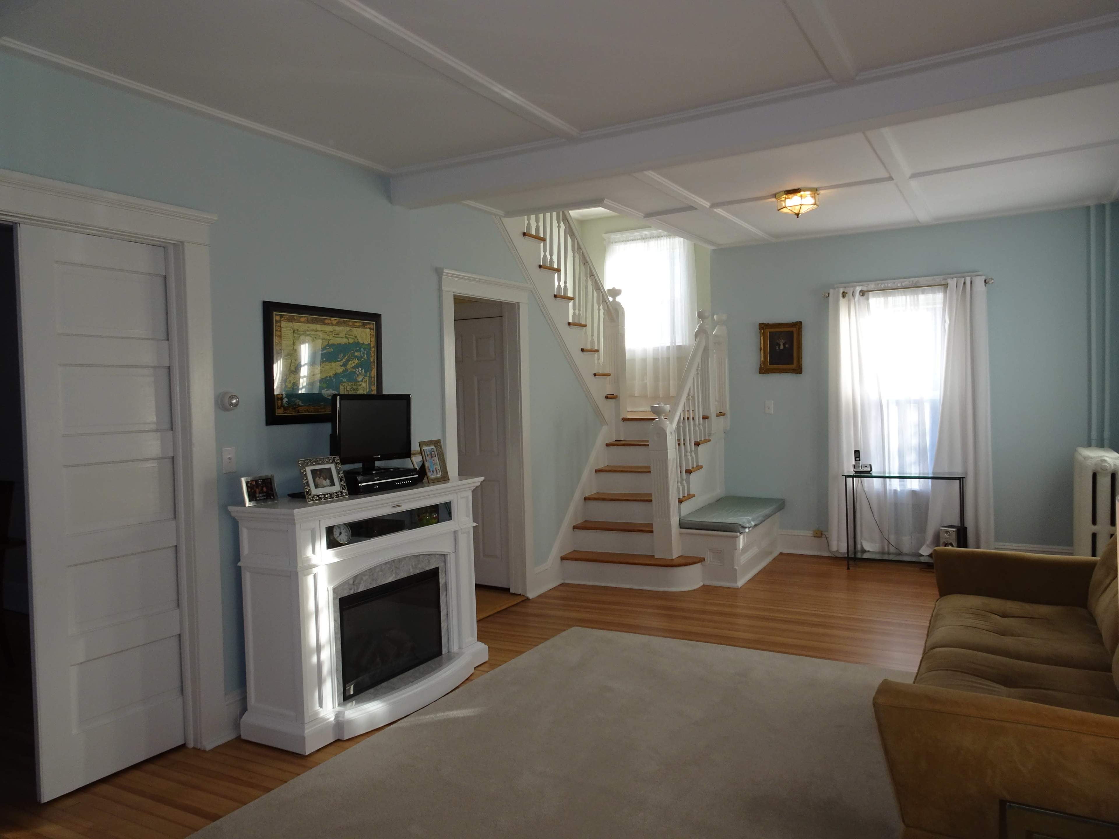 A softly lit living room with a staircase in the background, a fireplace with a television on top, and a beige couch facing a light-colored carpet.