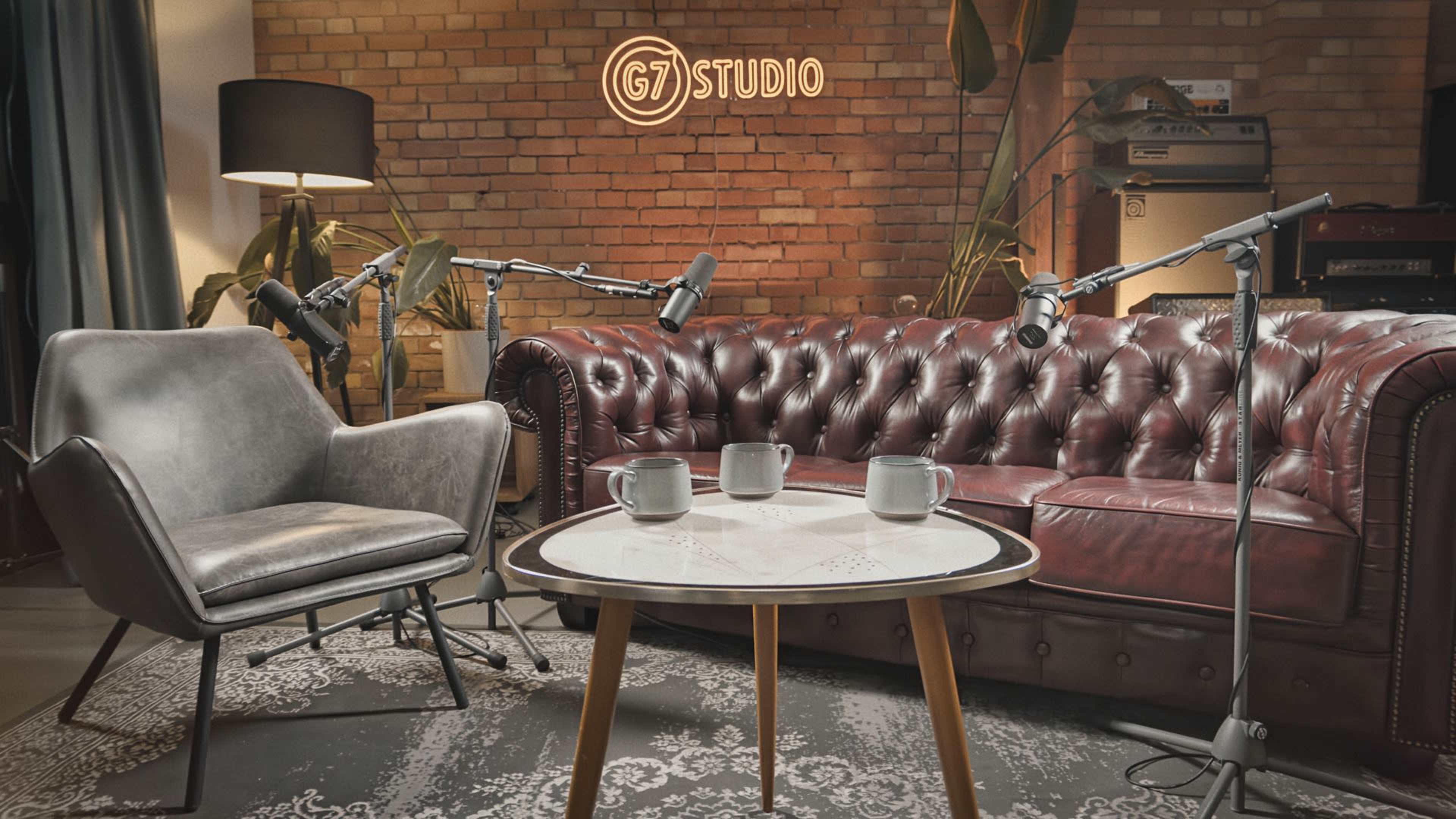 A cozy recording studio features a brown leather chesterfield sofa, two microphones on stands, a coffee table with three cups, and a brick wall with a "G7 STUDIO" sign.
