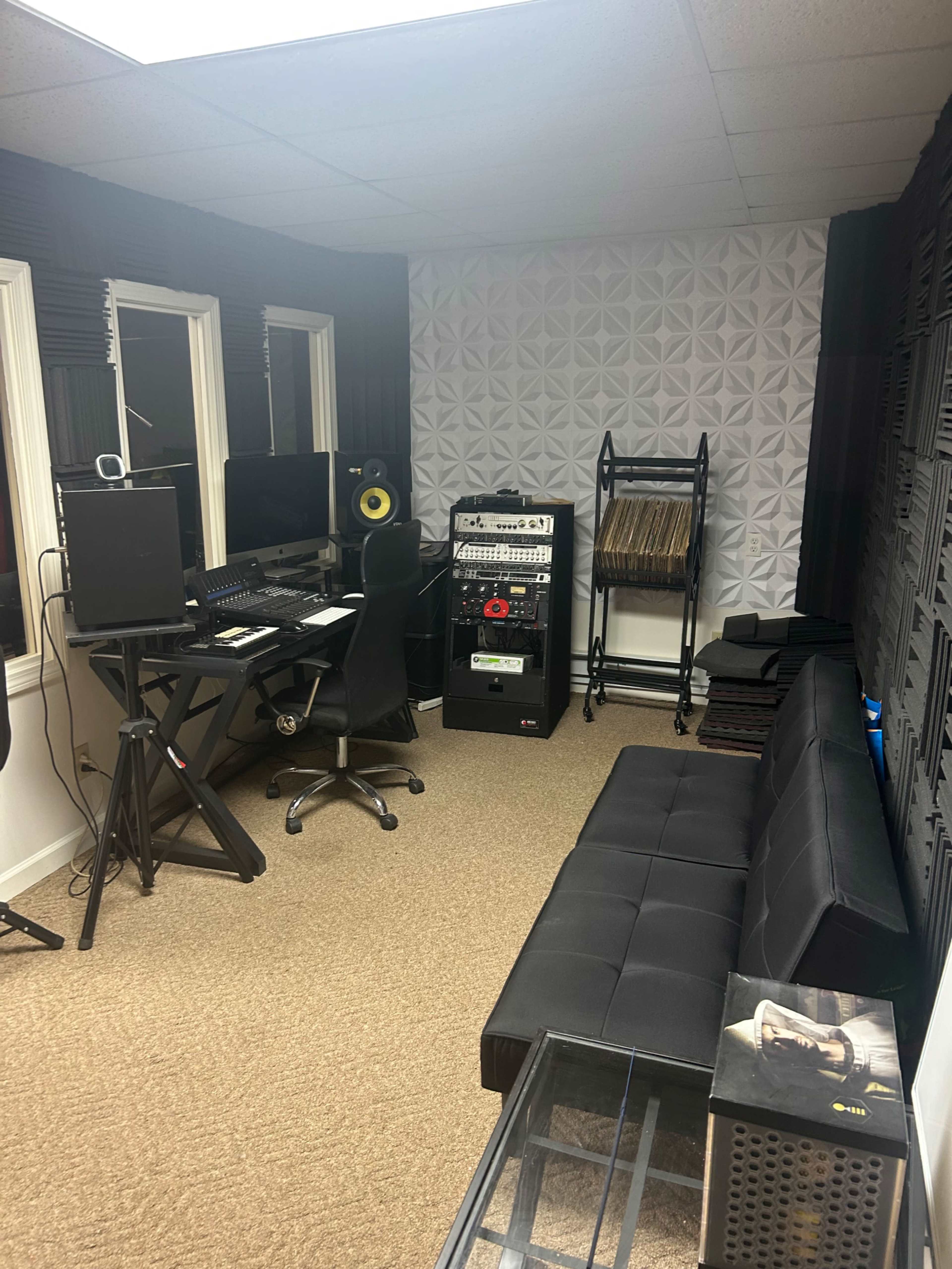 A small recording studio with a computer workstation, a black sofa, and audio equipment arranged against the walls.