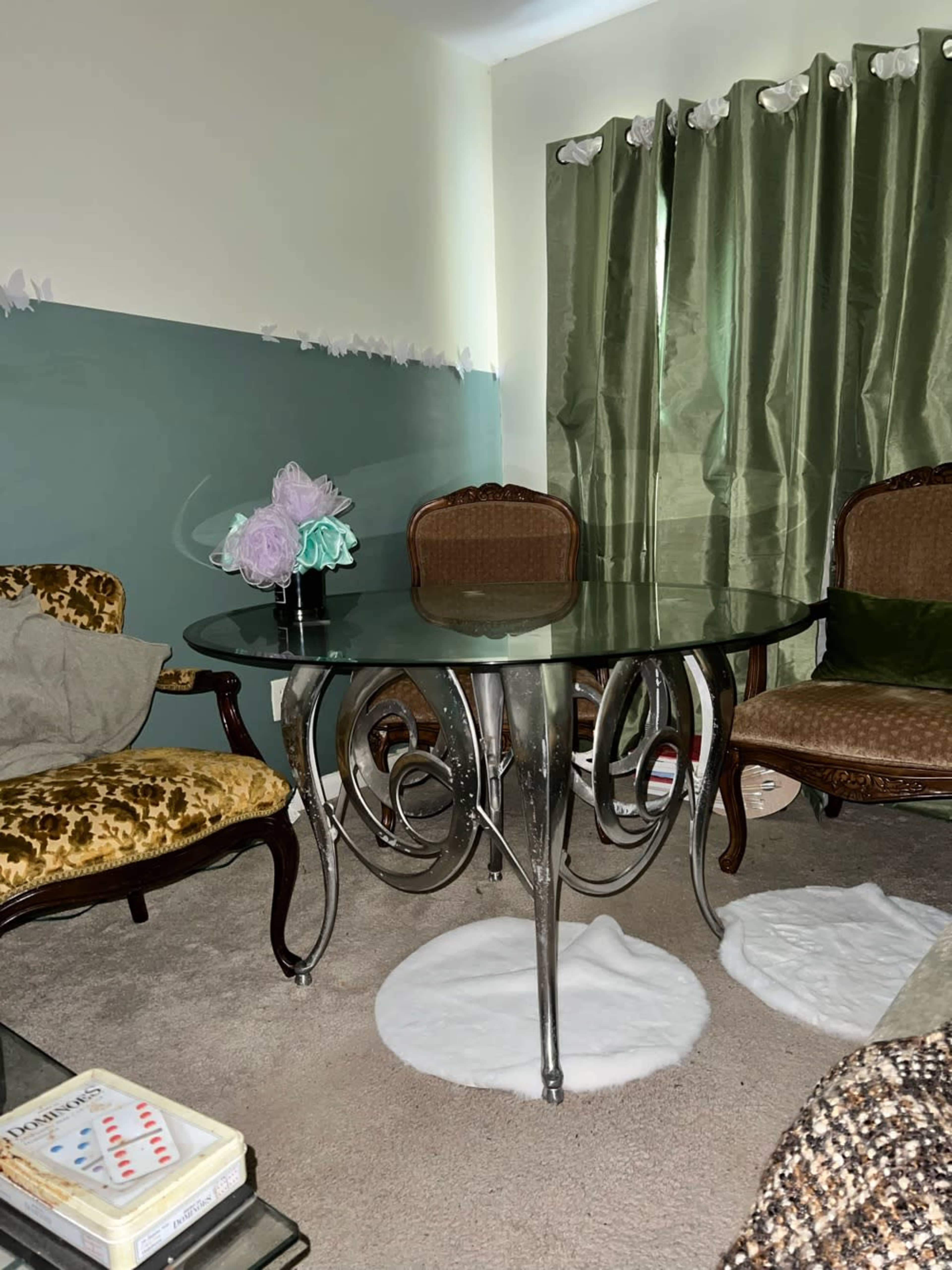 The image shows a glass-top table surrounded by ornate chairs in a room with green curtains and a decorative wall.