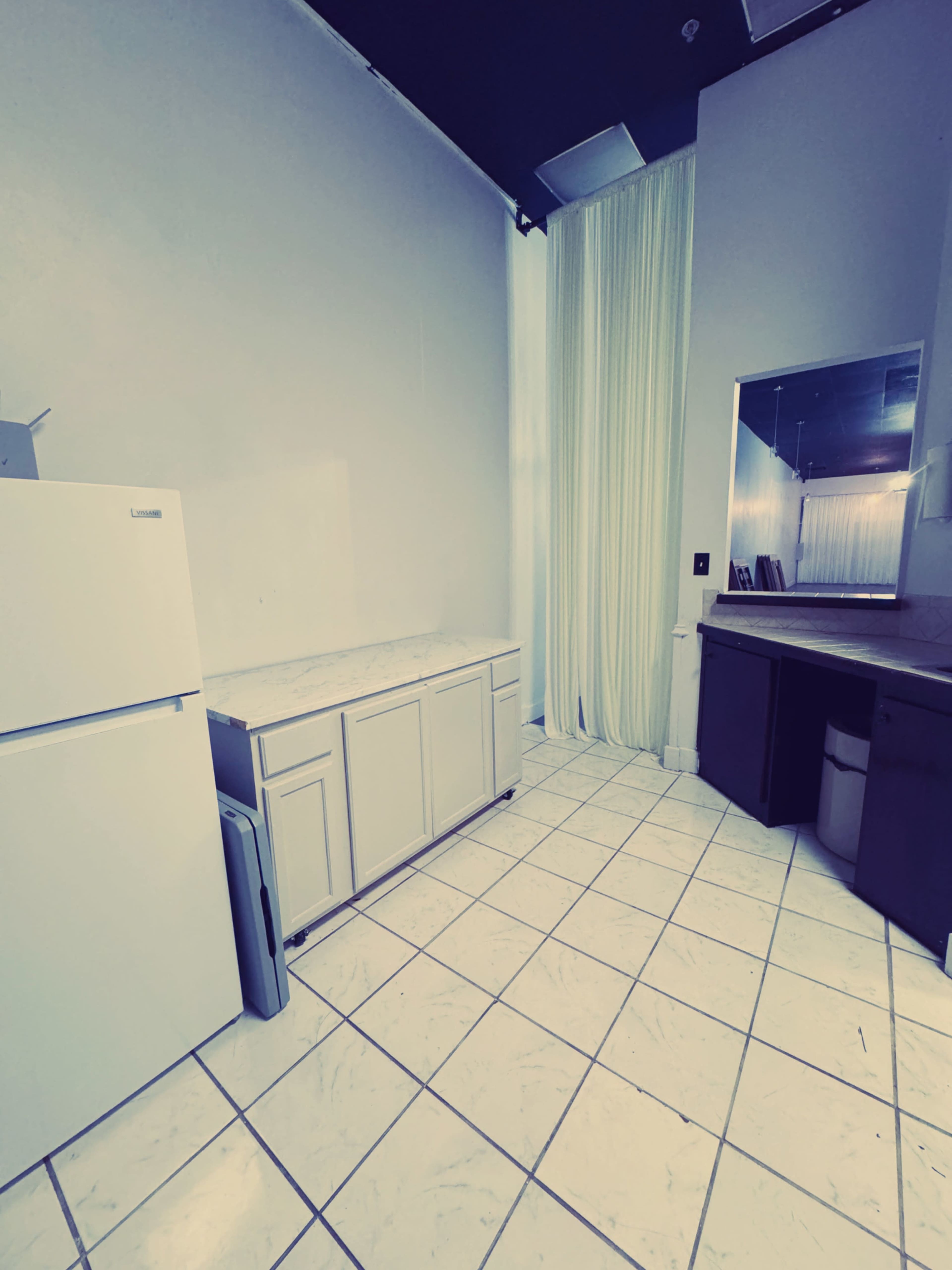 The image shows a small, minimally furnished kitchen area with a white refrigerator, a countertop with cabinets below, and a mirror on the wall.
