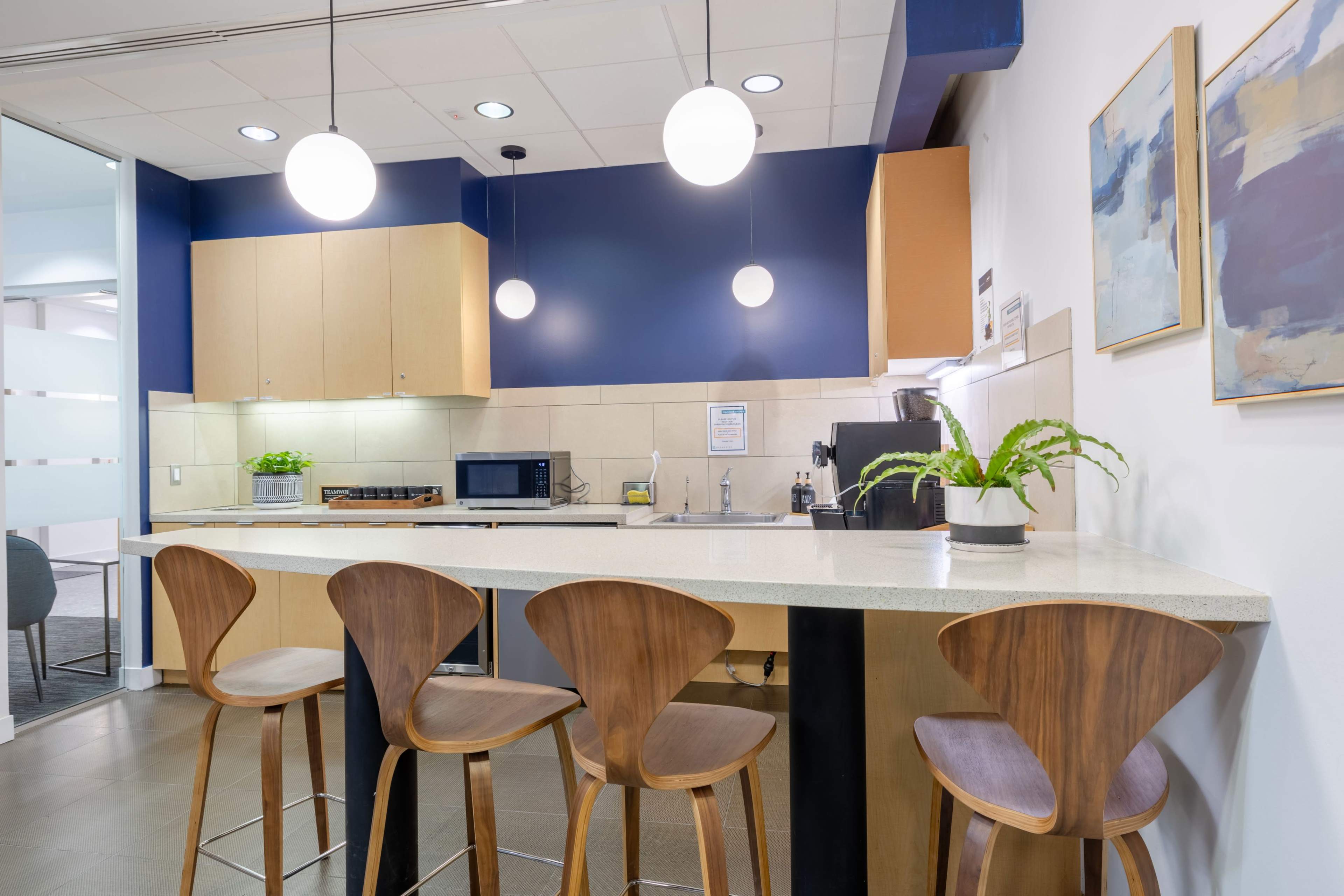 The image shows a modern kitchen area with a countertop and four wooden bar stools, equipped with a microwave, coffee maker, and potted plants against a backdrop of blue and light wood cabinetry.