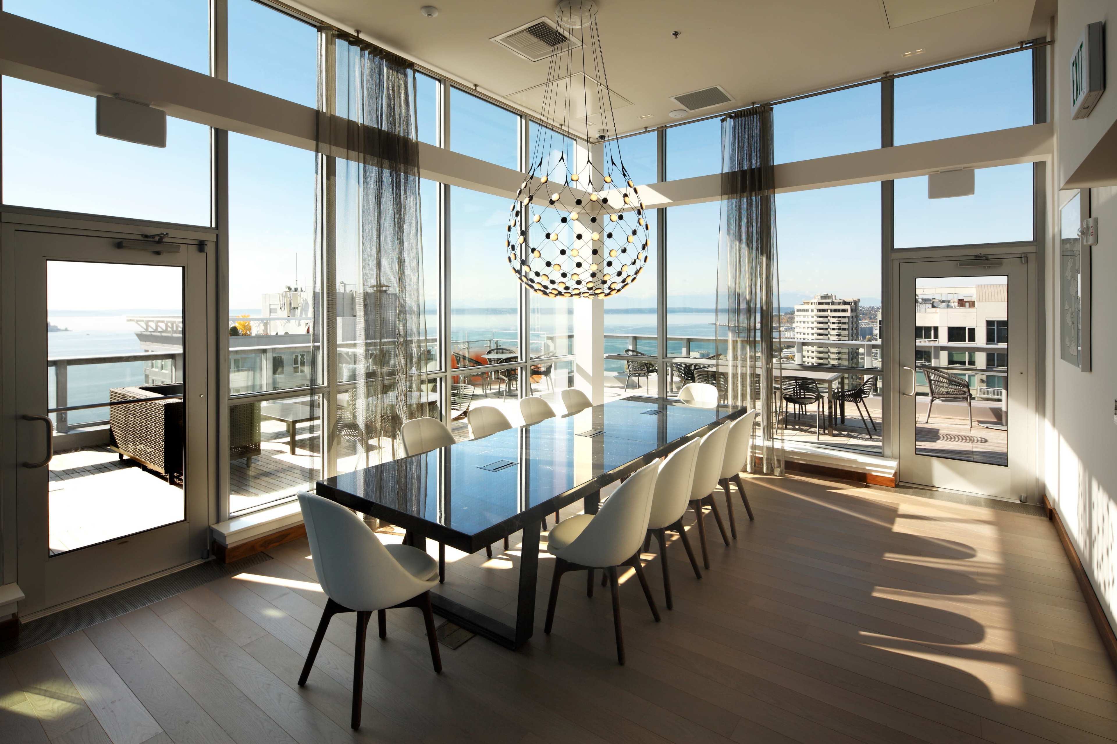 A modern dining area features a long table surrounded by white chairs, with large windows offering a view of the water and city skyline.
