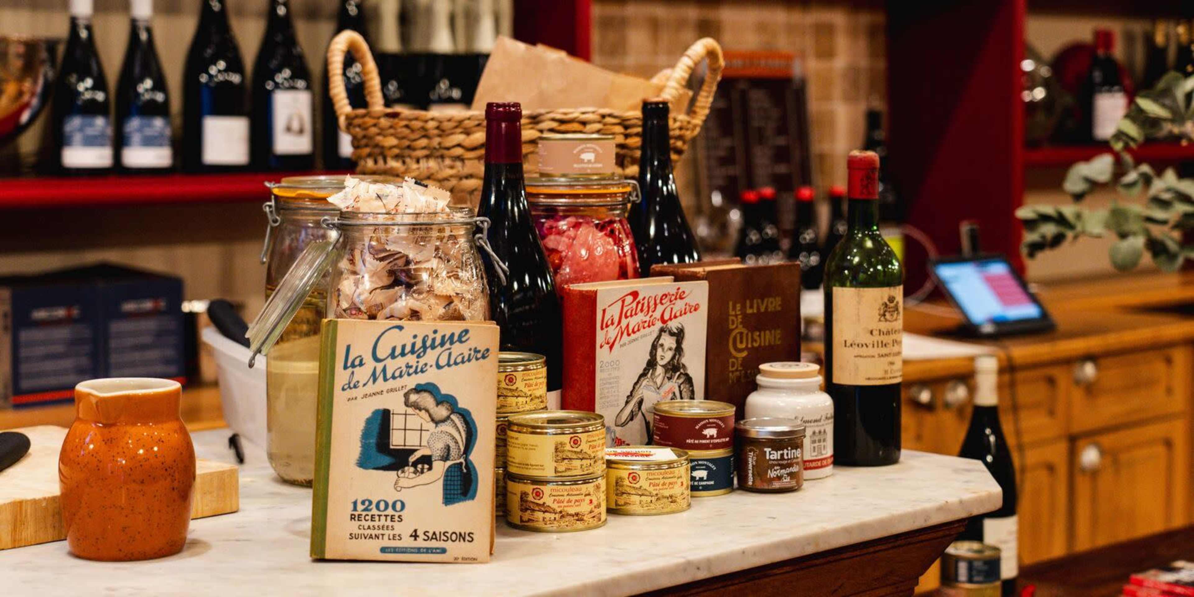 The image shows a countertop filled with various food items, including jars, bottles of wine, and canned products, all arranged alongside a decorative basket.