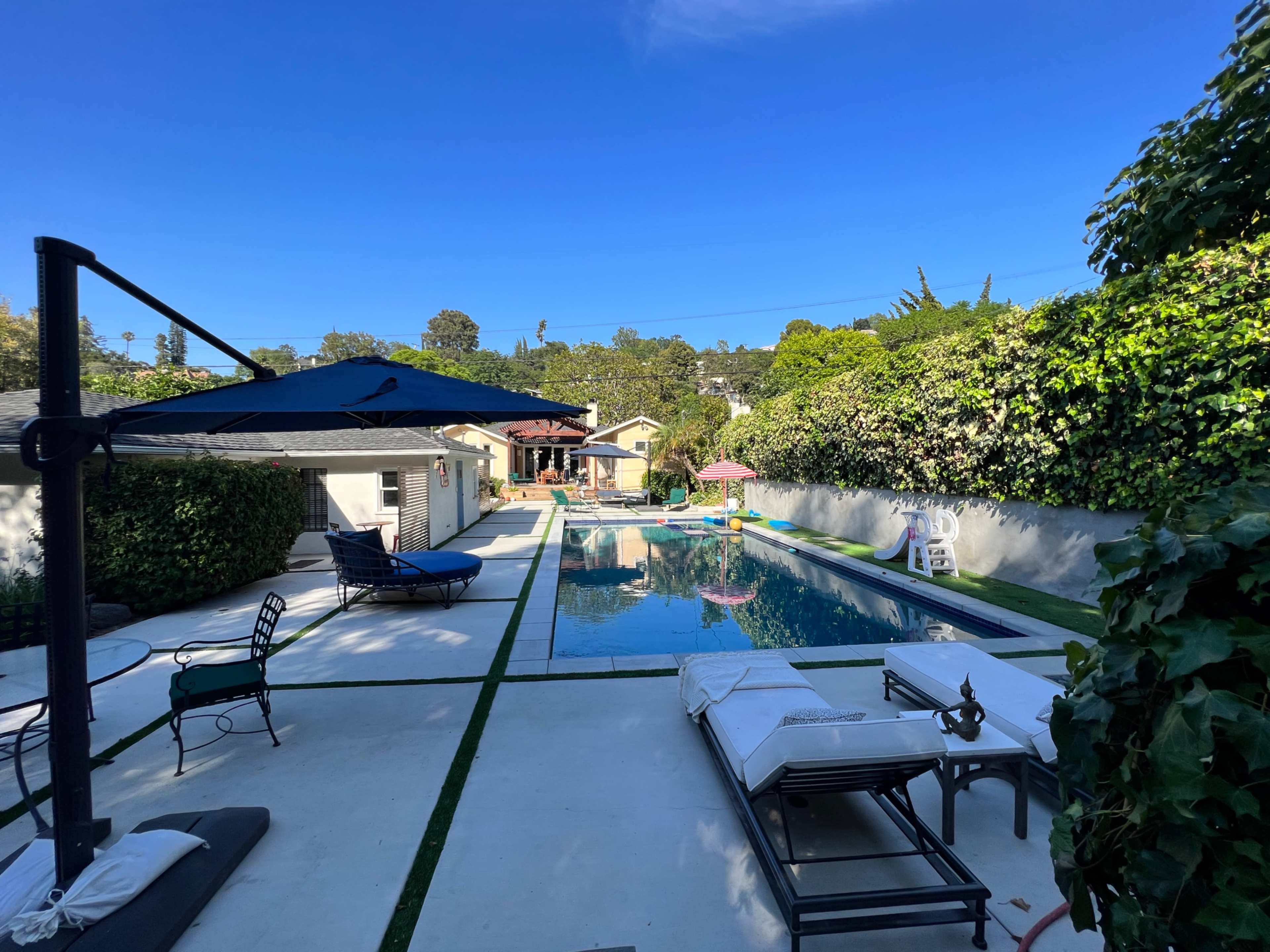 A backyard pool area with sun loungers, a large umbrella, and houses in the background under a clear blue sky.