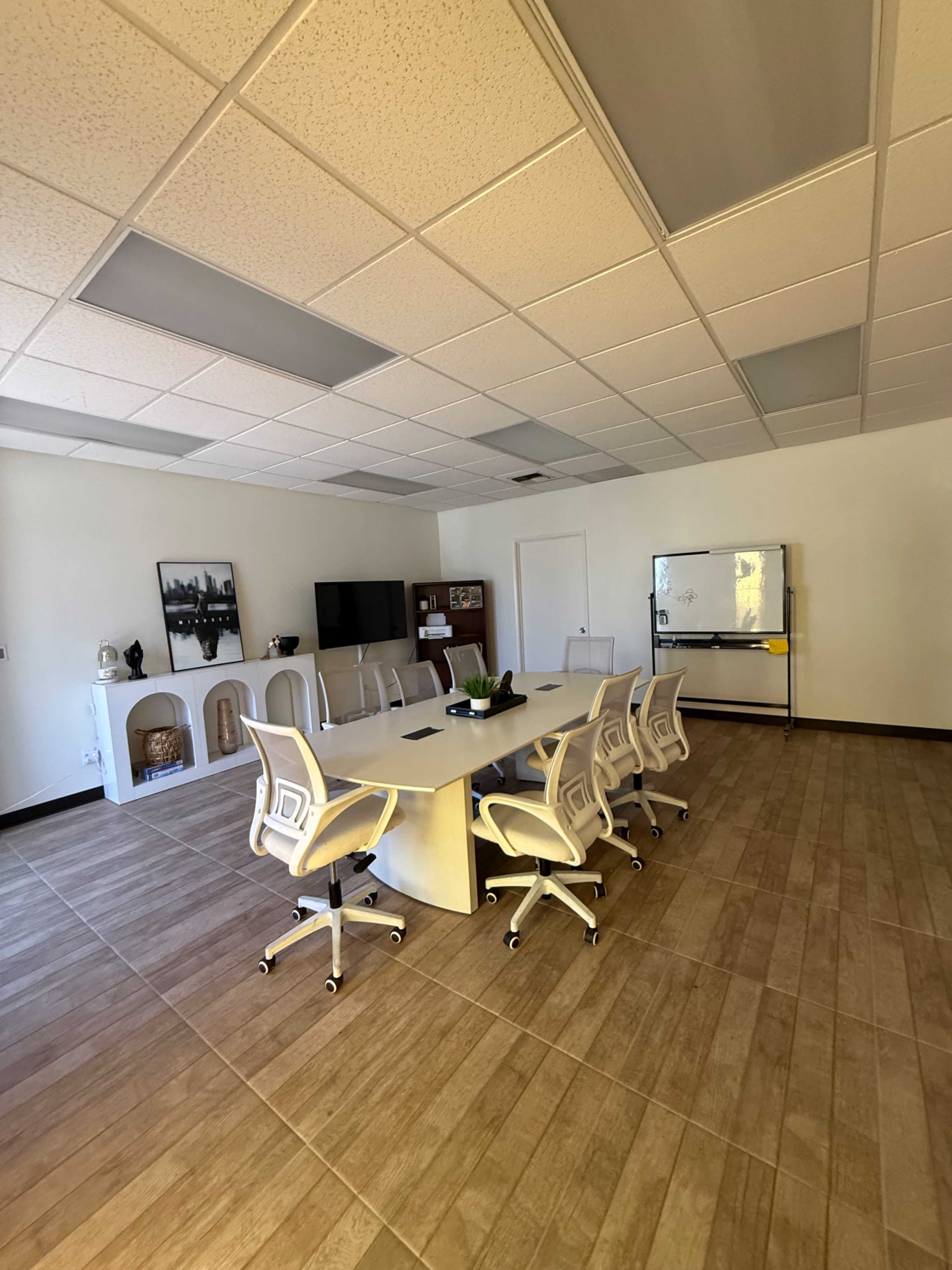 A modern conference room features a large white table surrounded by ergonomic chairs, with a television and whiteboard visible on the walls.