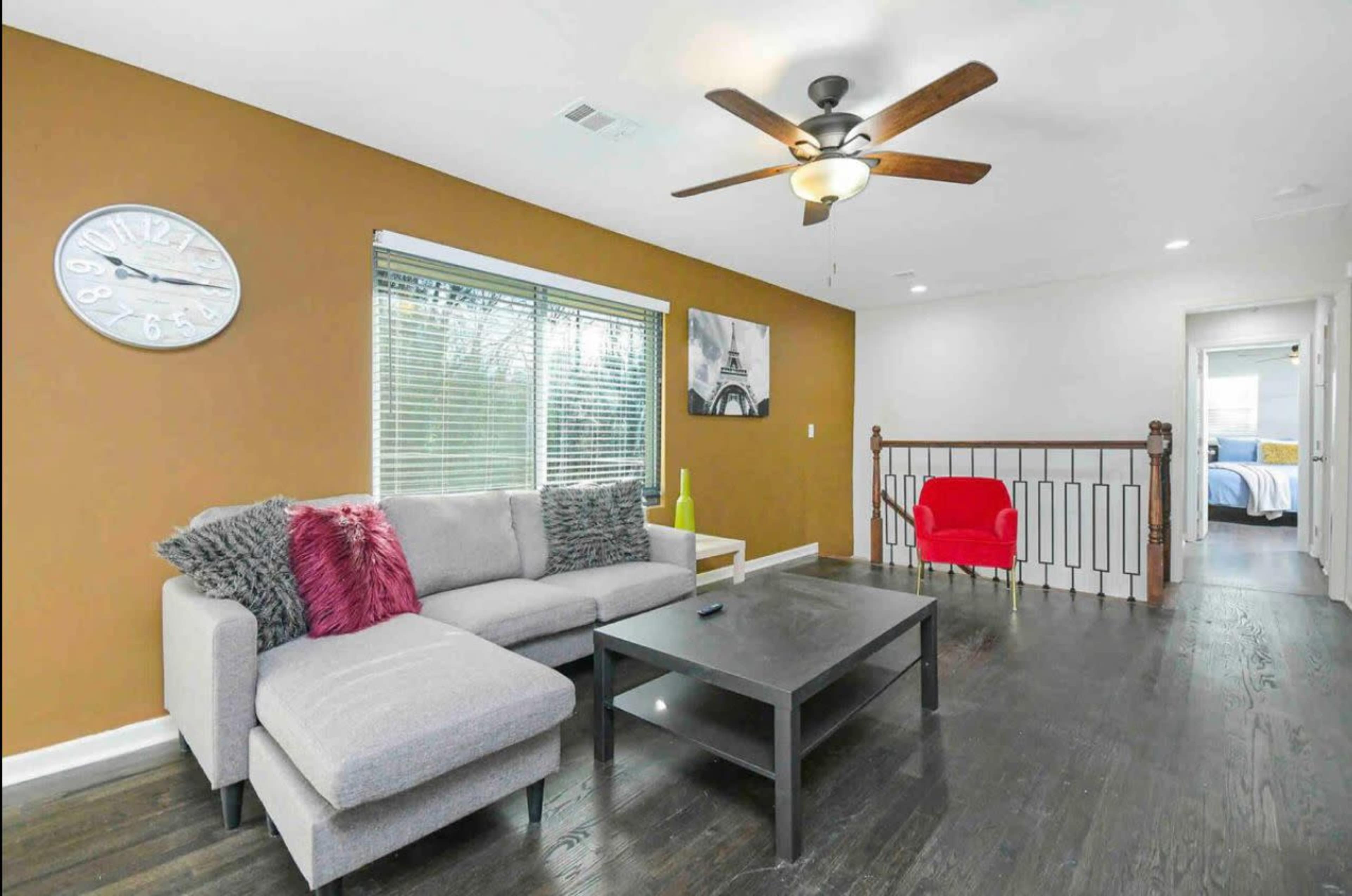 The image shows a living room with a gray sectional sofa, a black coffee table, and a ceiling fan, featuring a large window with blinds and a view of a nearby staircase.