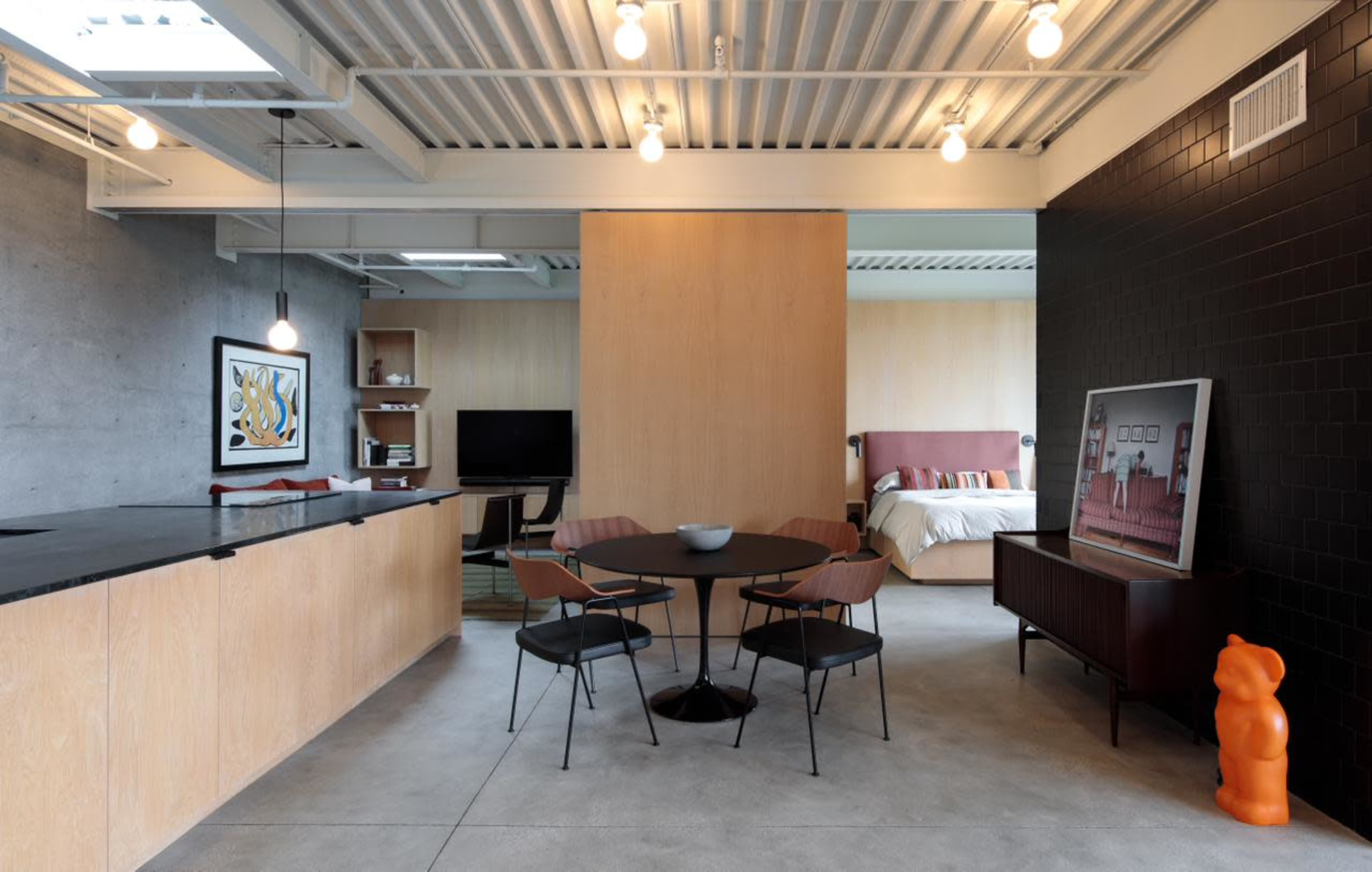 The image shows a modern open-concept living space featuring a kitchen area with a black countertop, a dining table with chairs, and a bedroom visible in the background.