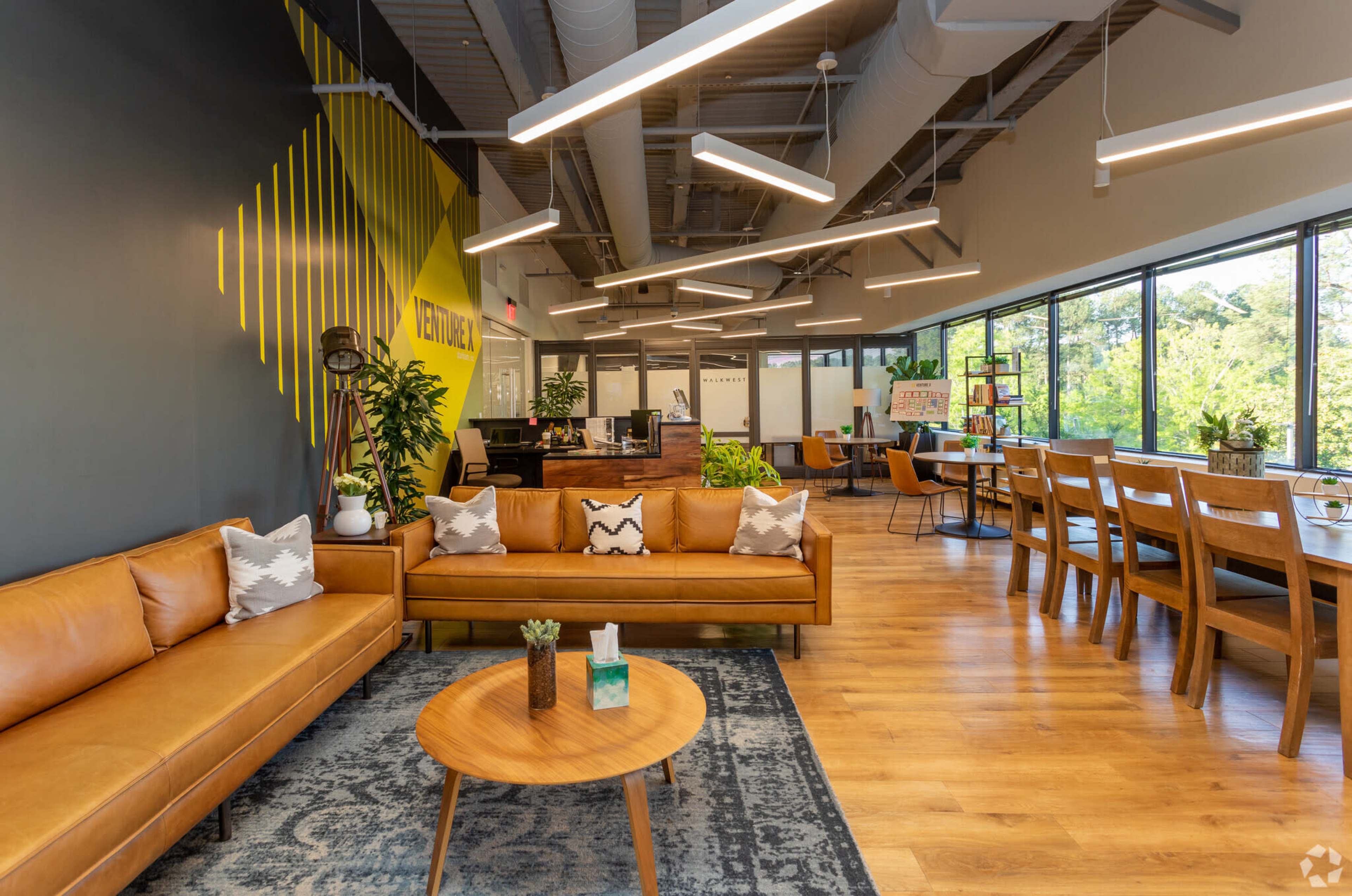The image shows a modern office interior featuring brown leather couches, a wooden coffee table, and large windows overlooking greenery.