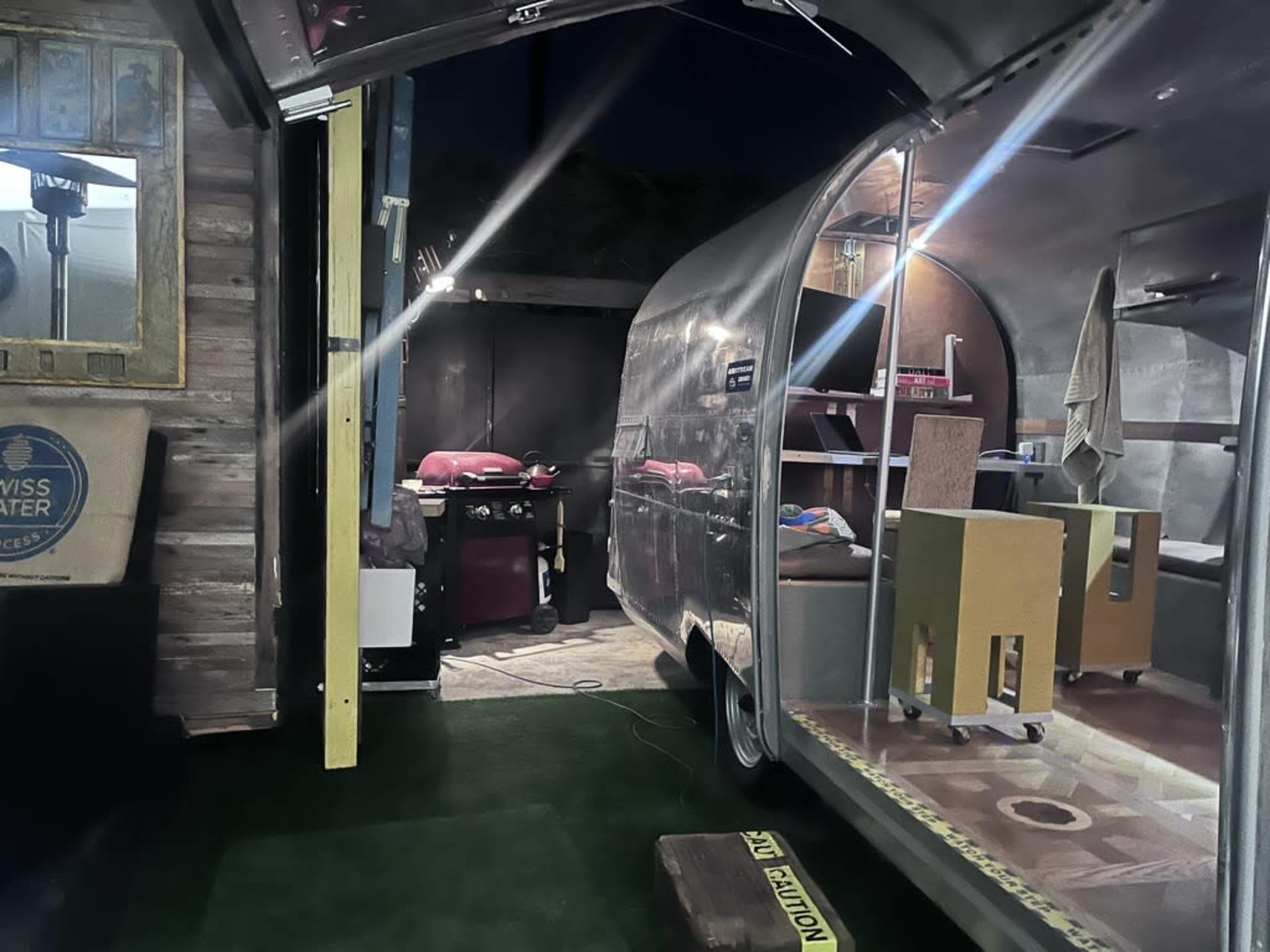 A dimly lit outdoor area featuring an Airstream trailer with an open door, revealing a cozy interior with furniture and kitchen equipment visible.