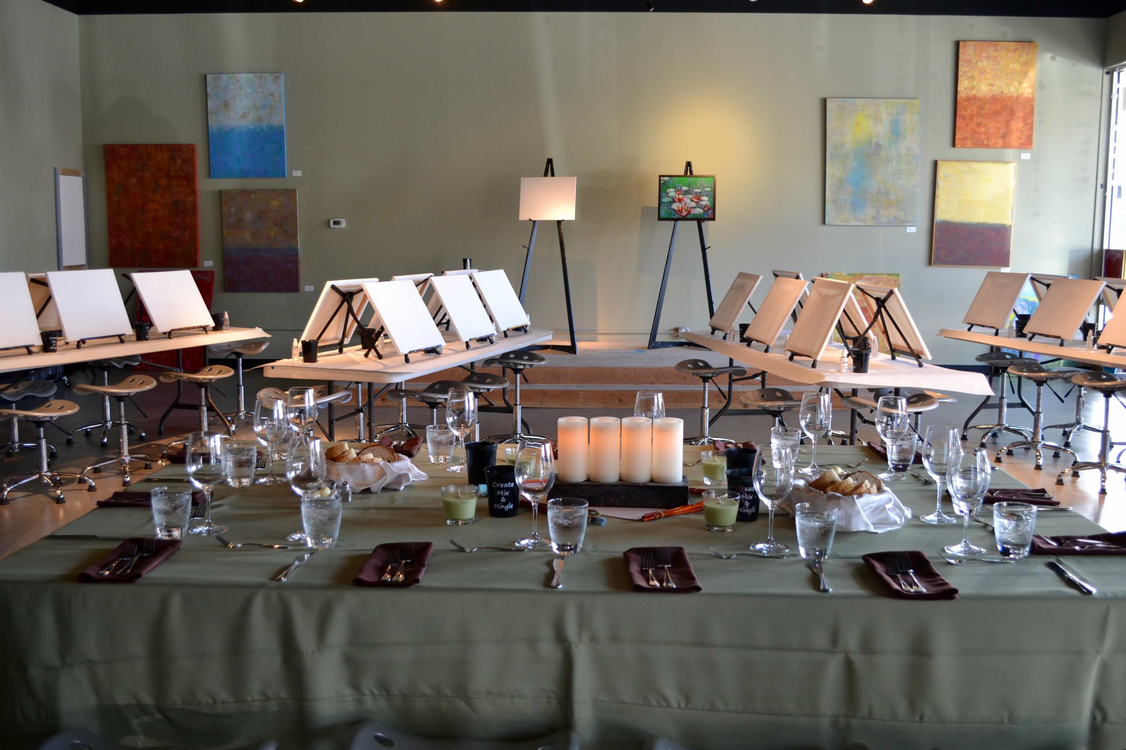 A spacious art studio features a long dining table set with glasses and plates, surrounded by easels with blank canvases.