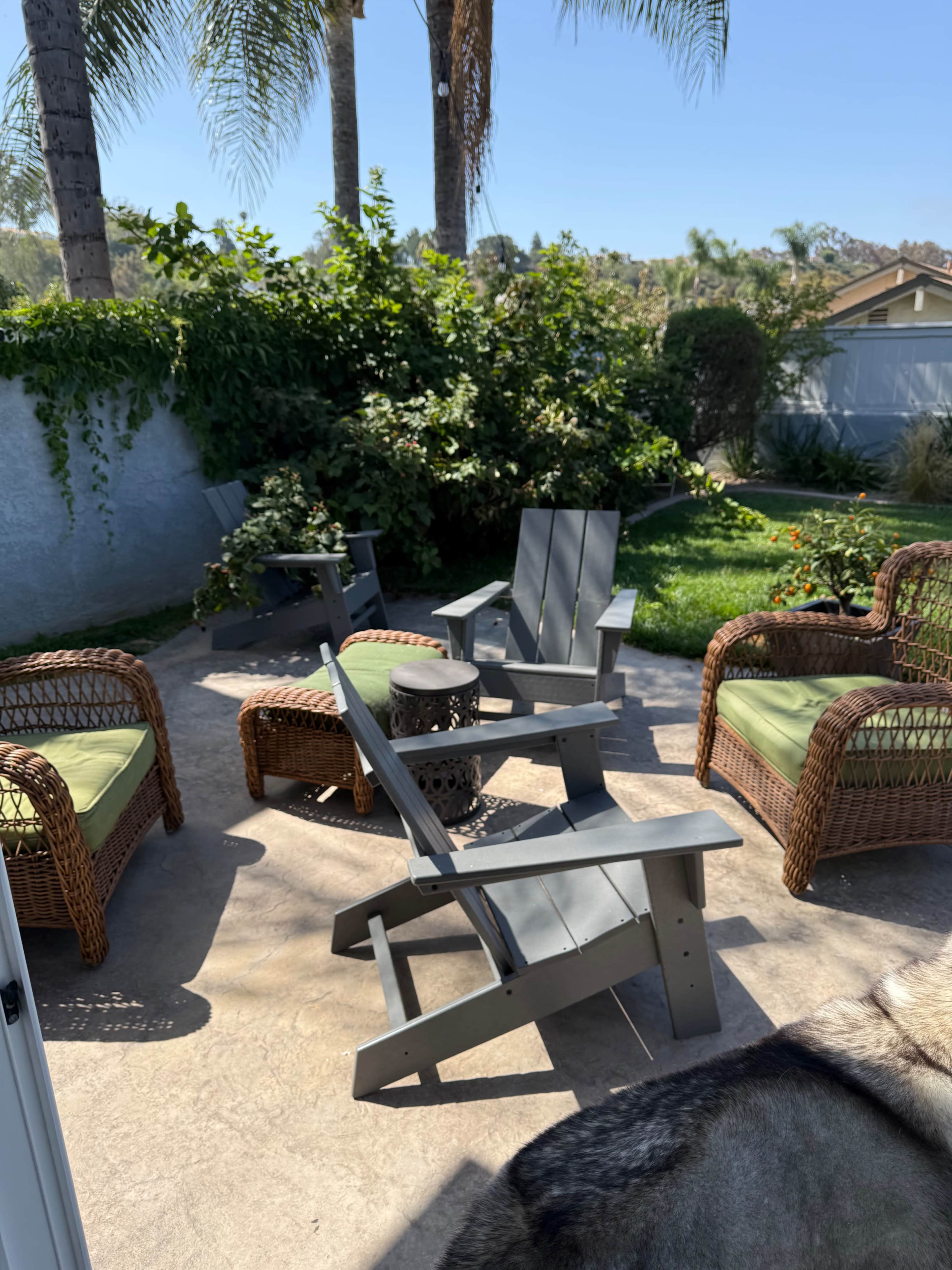 The image shows a patio area with four outdoor chairs, two of which are Adirondack-style, surrounded by greenery and a small round table.