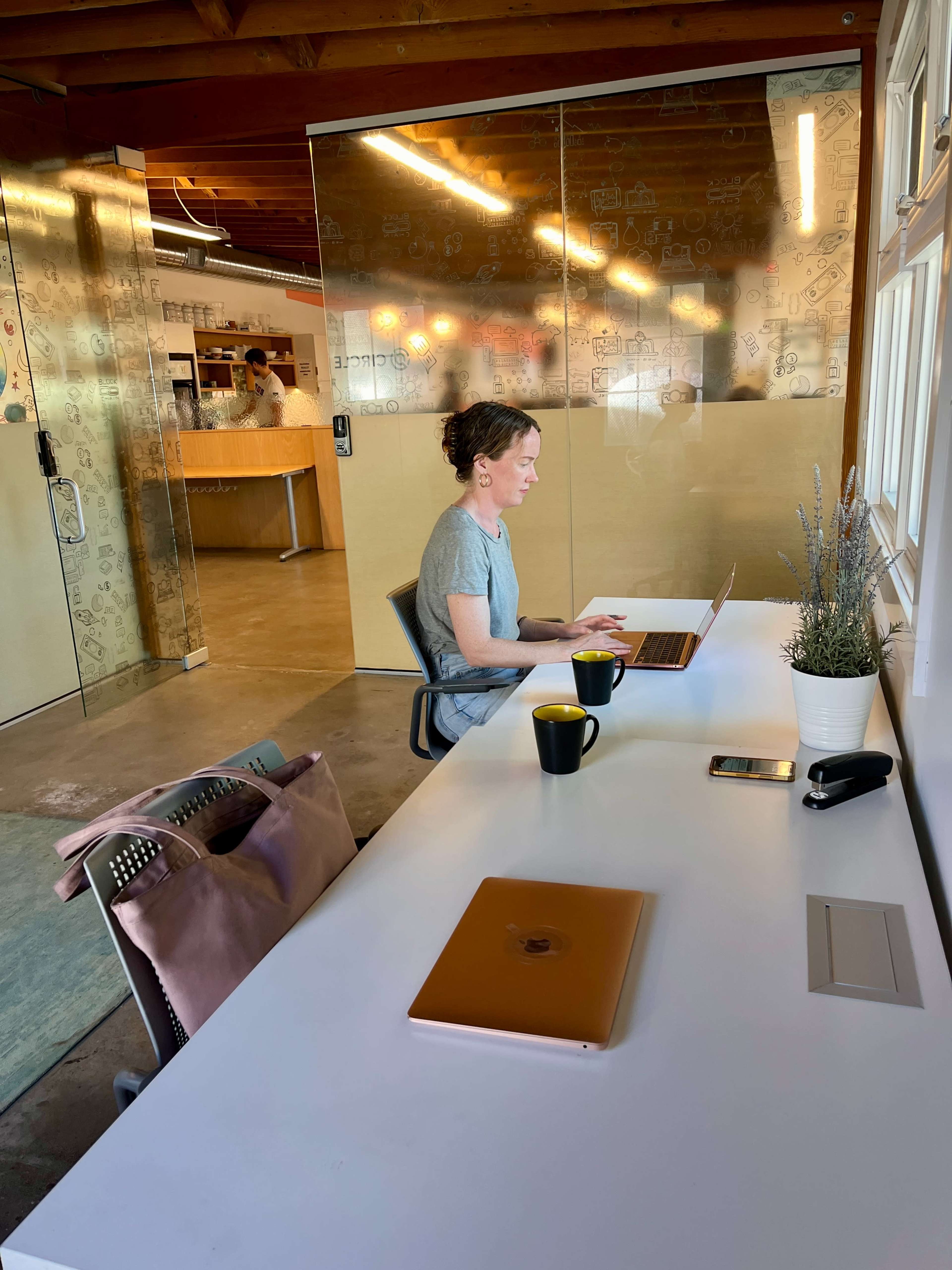 A woman works on a laptop at a desk in a modern office space with large glass panels and a kitchen area in the background.