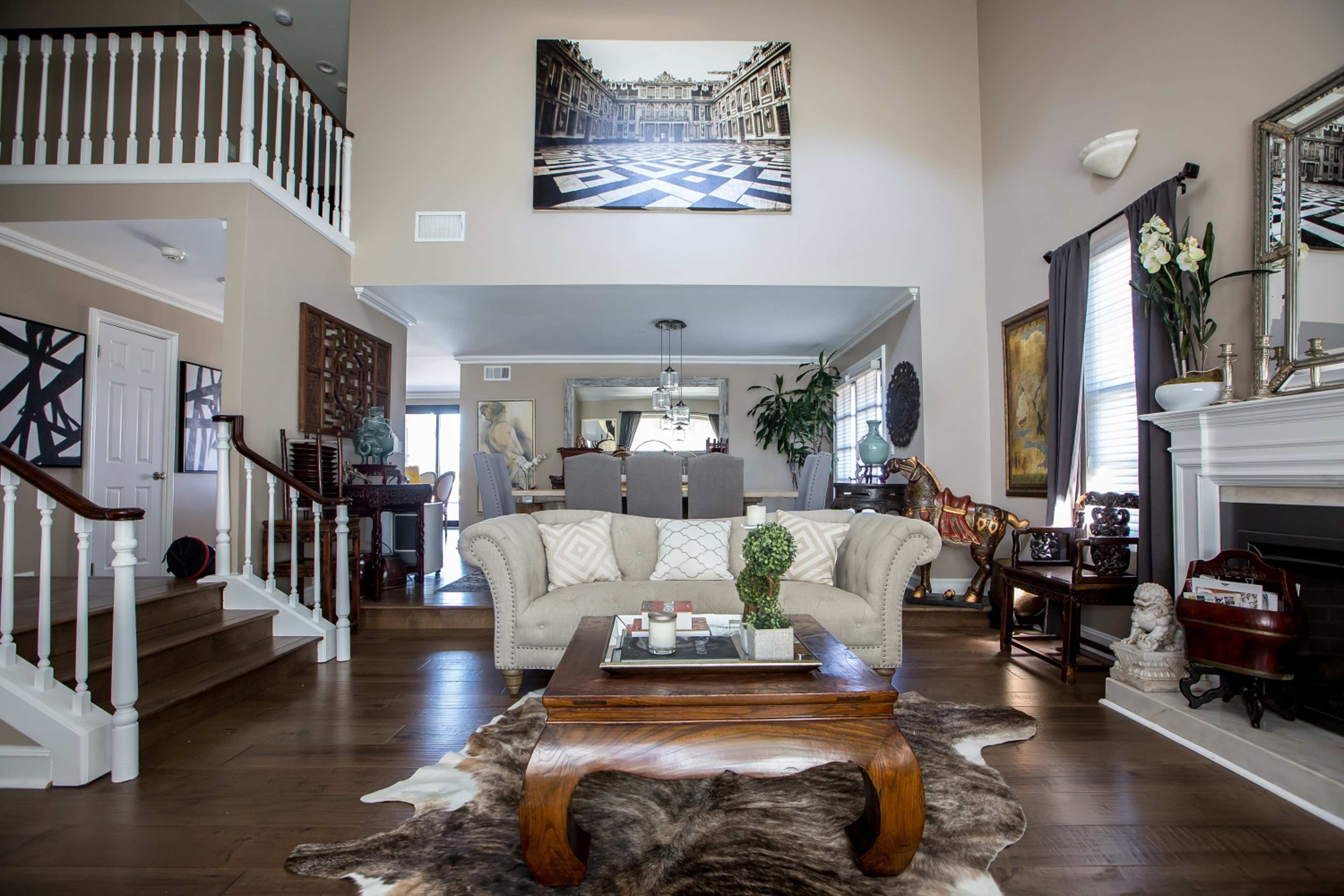 The image depicts a spacious living room featuring a beige sofa, a wooden coffee table on a cowhide rug, and elegant decor elements, illuminated by natural light from large windows.