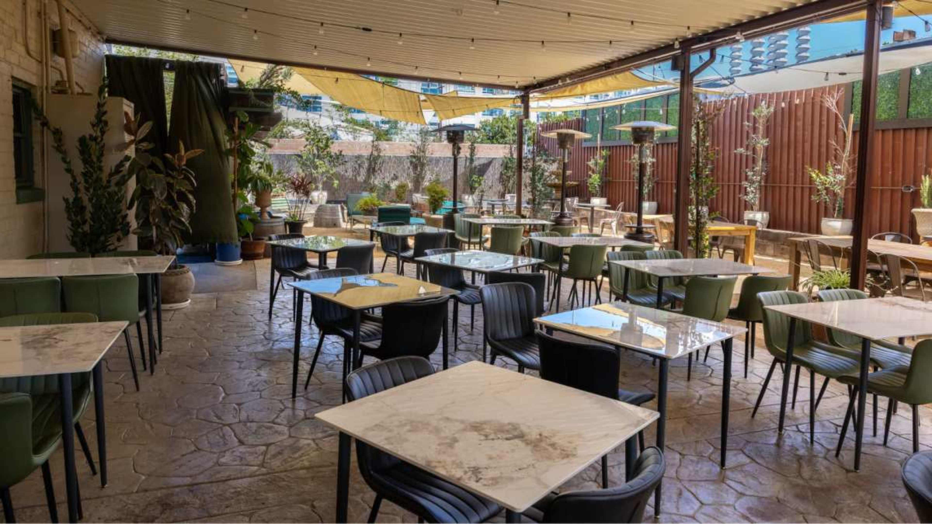 A spacious outdoor dining area features numerous tables with marble tops and green and black chairs, surrounded by potted plants and a canopy overhead.