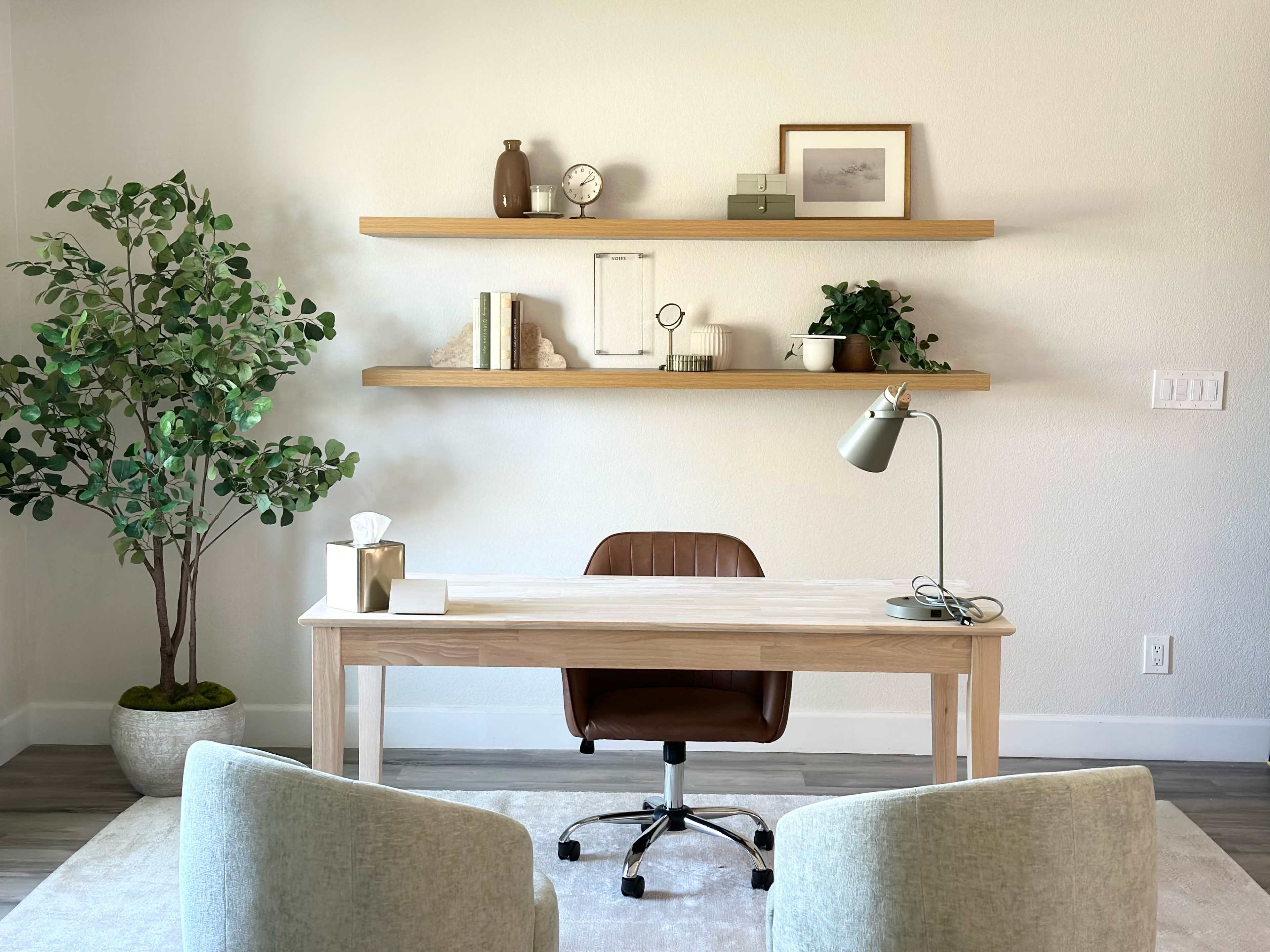 A minimalist home office features a wooden desk with an ergonomic chair, flanked by two upholstered chairs and adorned with shelves displaying various decorative items.