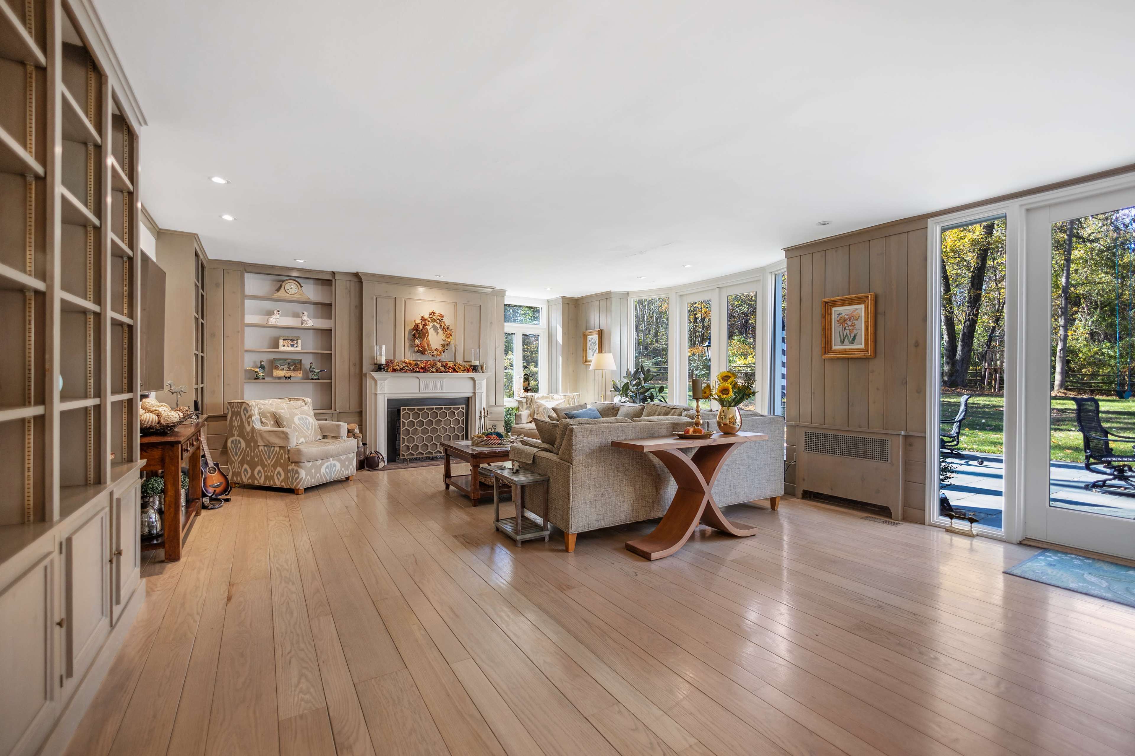 A bright living room features large windows, a fireplace, and a seating area with light-colored furniture and wooden flooring.