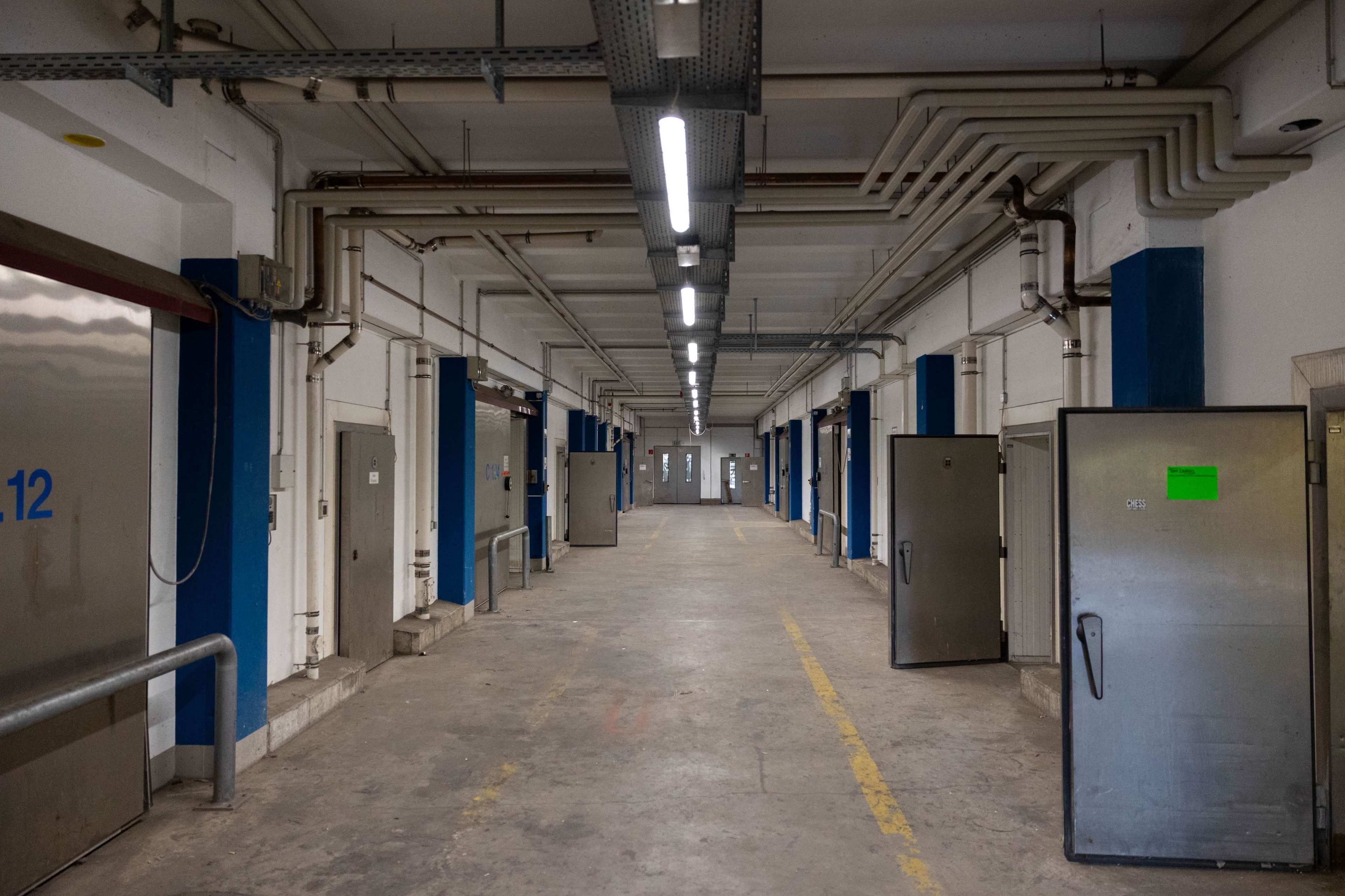 The image shows a long, empty corridor in a building with metal doors on either side and pipes running along the ceiling.