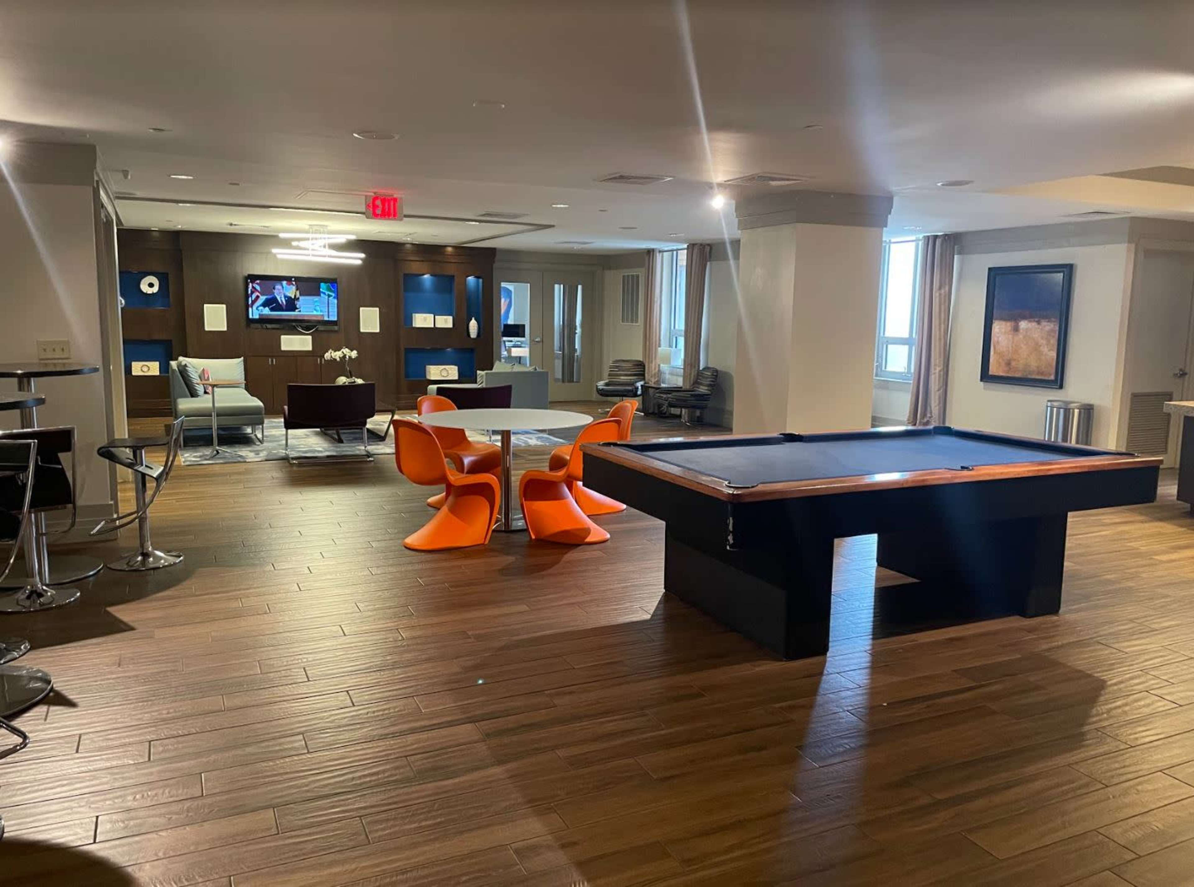 A modern communal lounge area featuring a billiards table, a round dining table with orange chairs, and a seating space with a television in the background.
