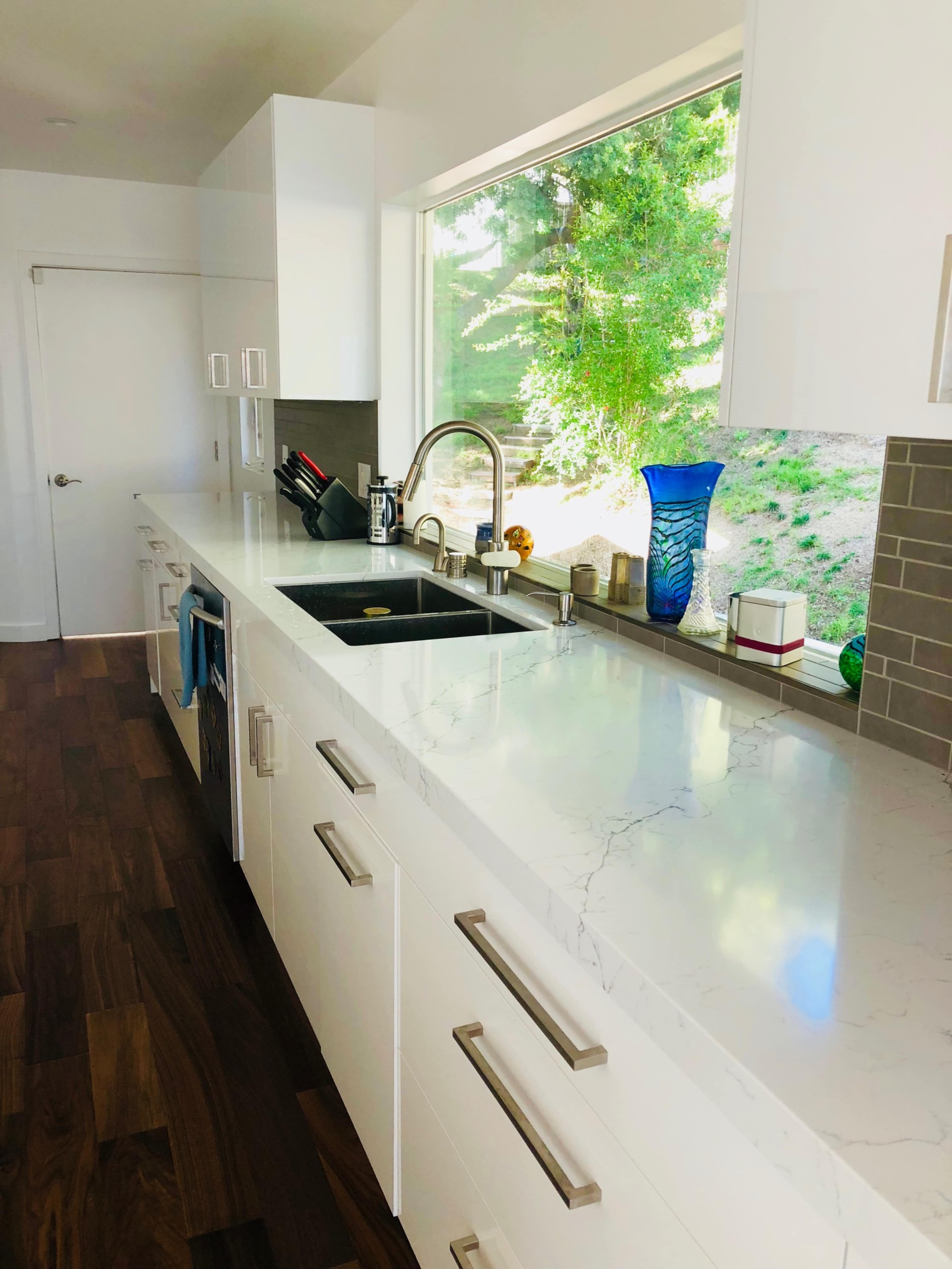 A modern kitchen features a white countertop with a sink and large window overlooking greenery.