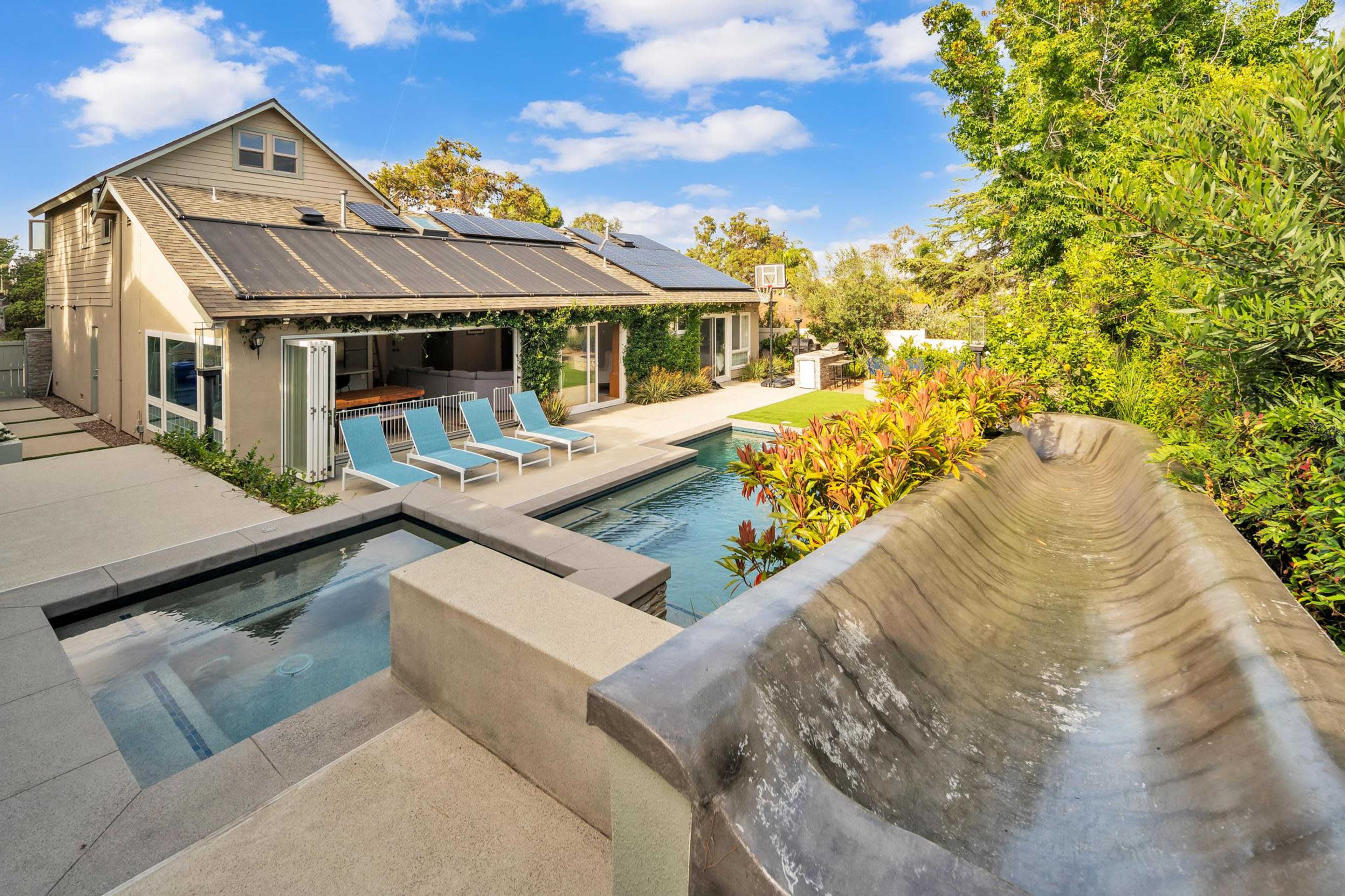 A modern backyard with a swimming pool, a water slide, and a house featuring solar panels and an outdoor seating area.